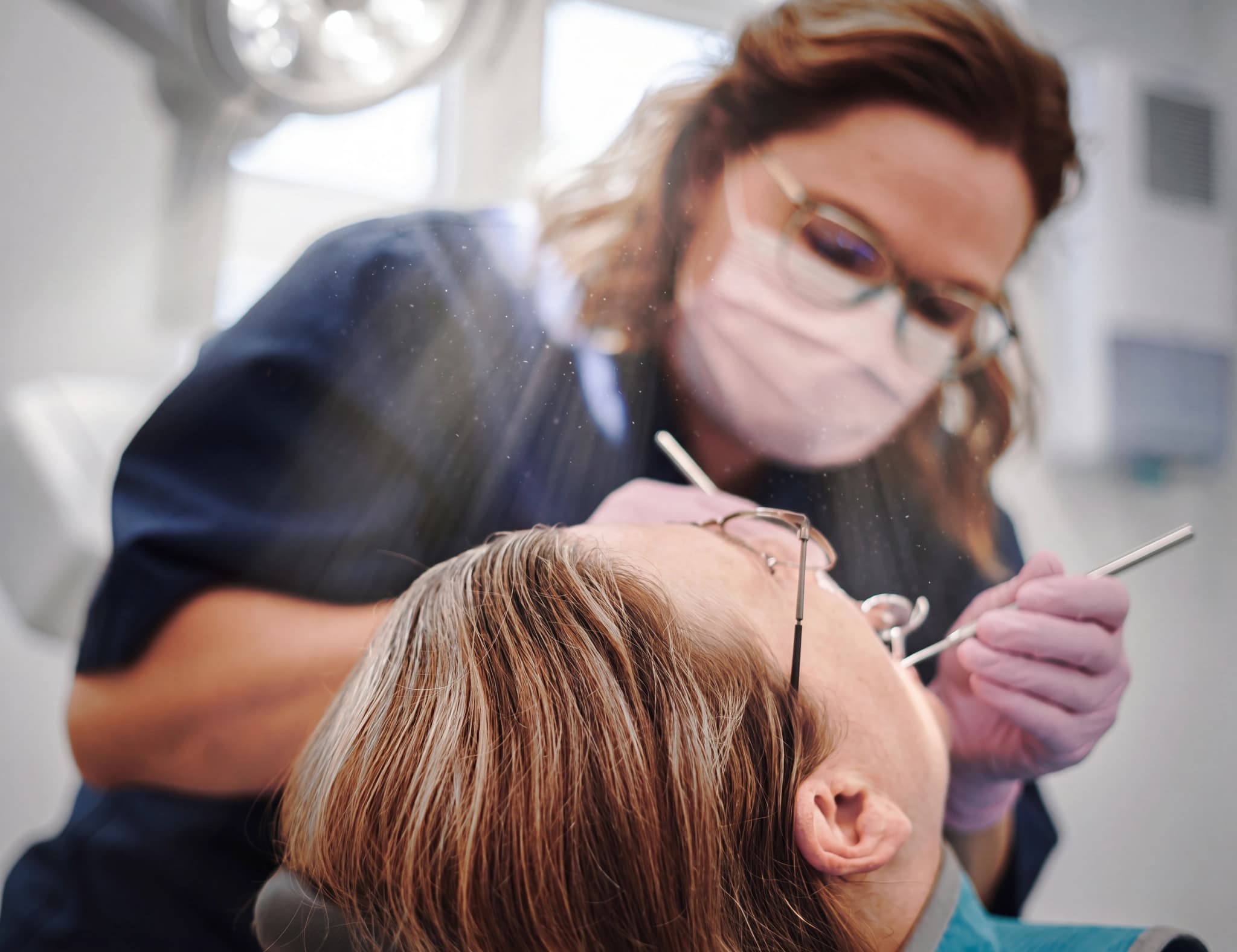Tandläkare med munskydd och glasögon undersöker en patients tänder med ett instrument.