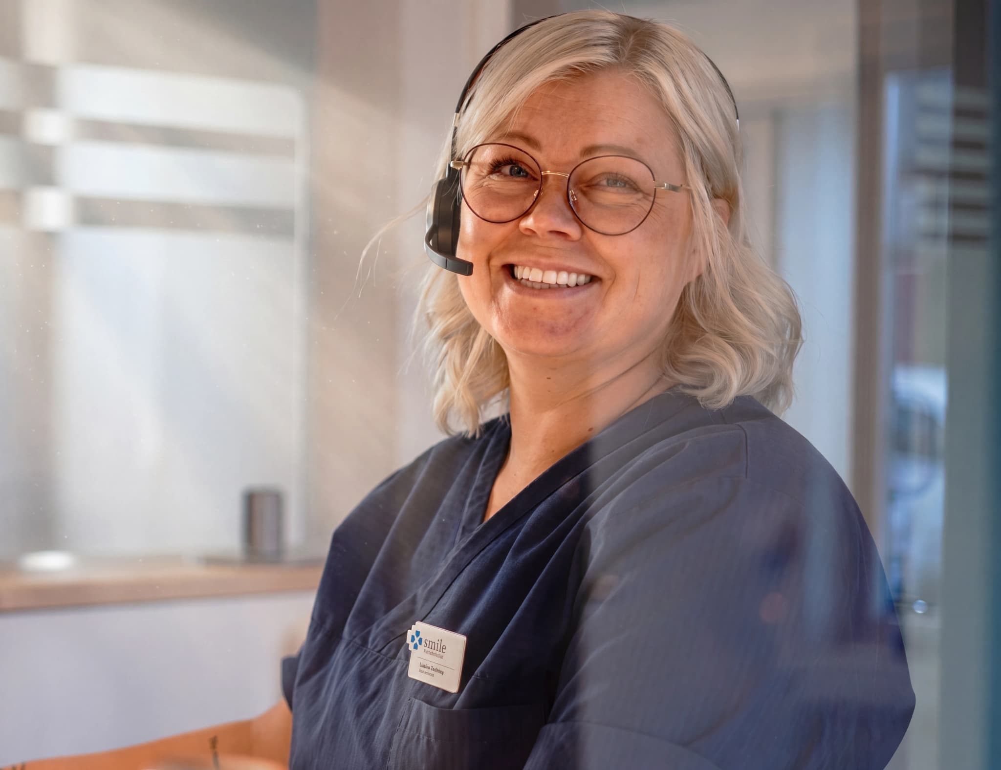 En kvinna med glasögon och headset ler mot kameran, klädd i en mörkblå arbetsklädsel med en namnskylt.