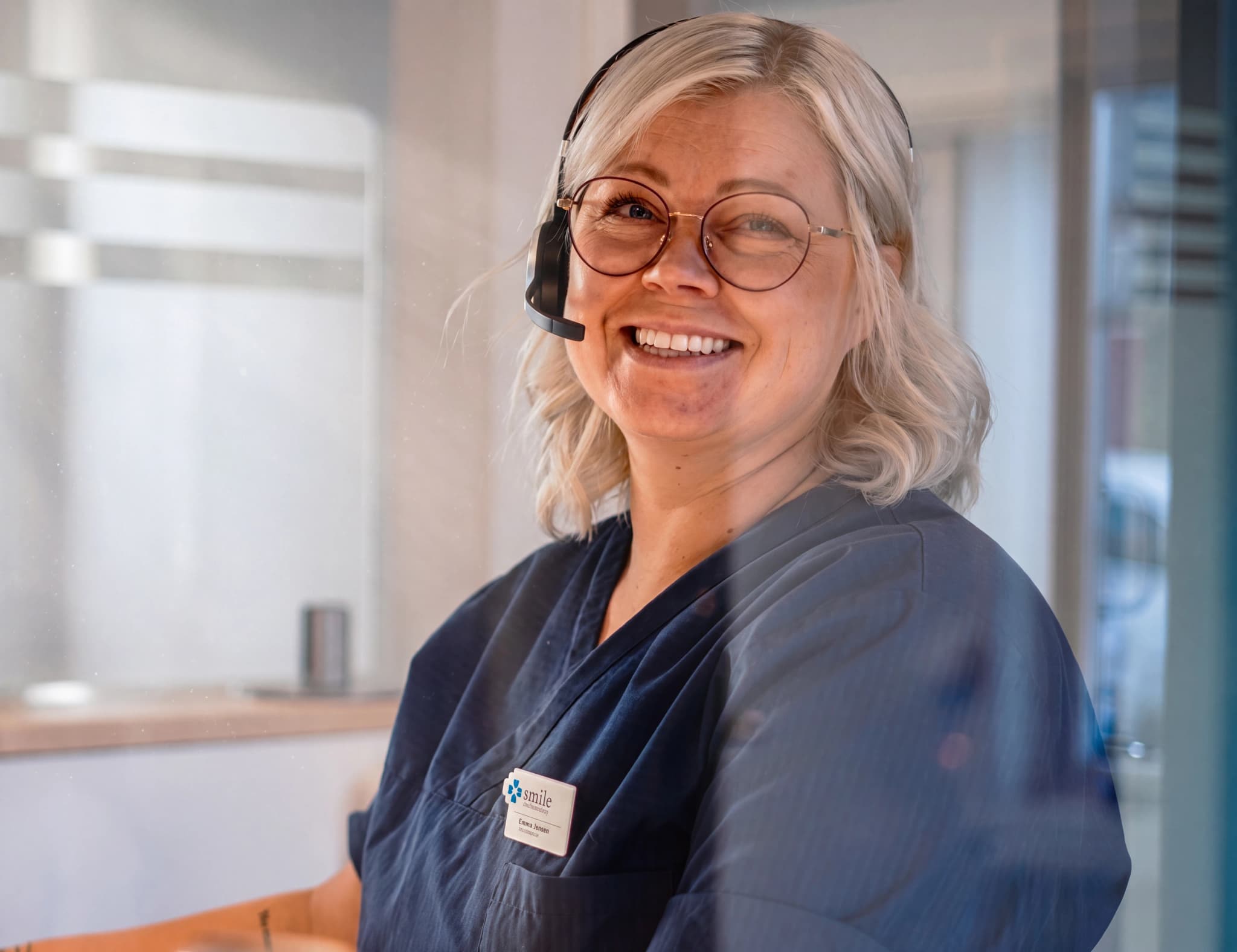 En kvinna med glasögon och headset ler mot kameran och sitter bakom ett skrivbord.
