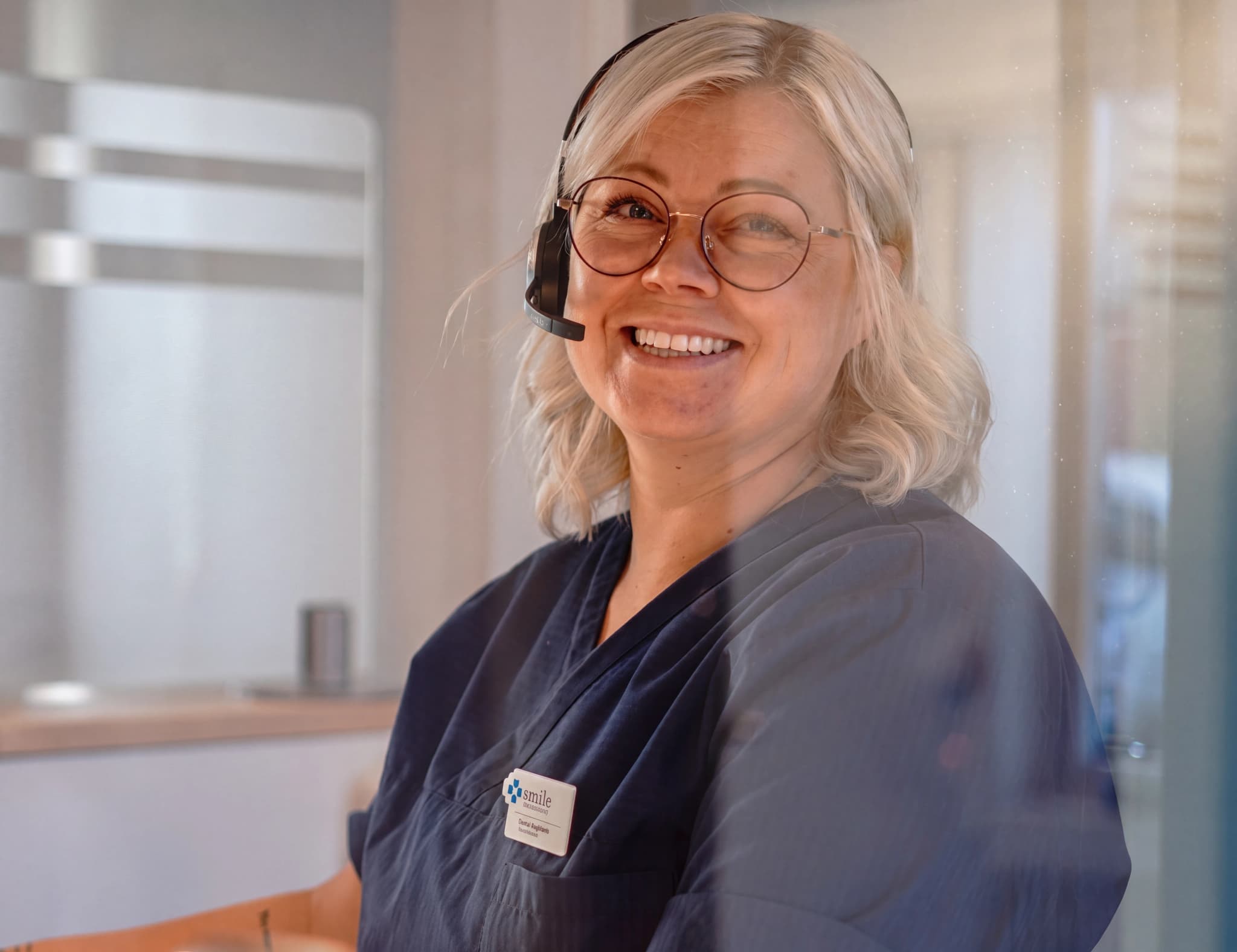 En kvinna med glasögon och headset ler mot kameran, klädd i mörkblå arbetskläder med ett namnskylt.