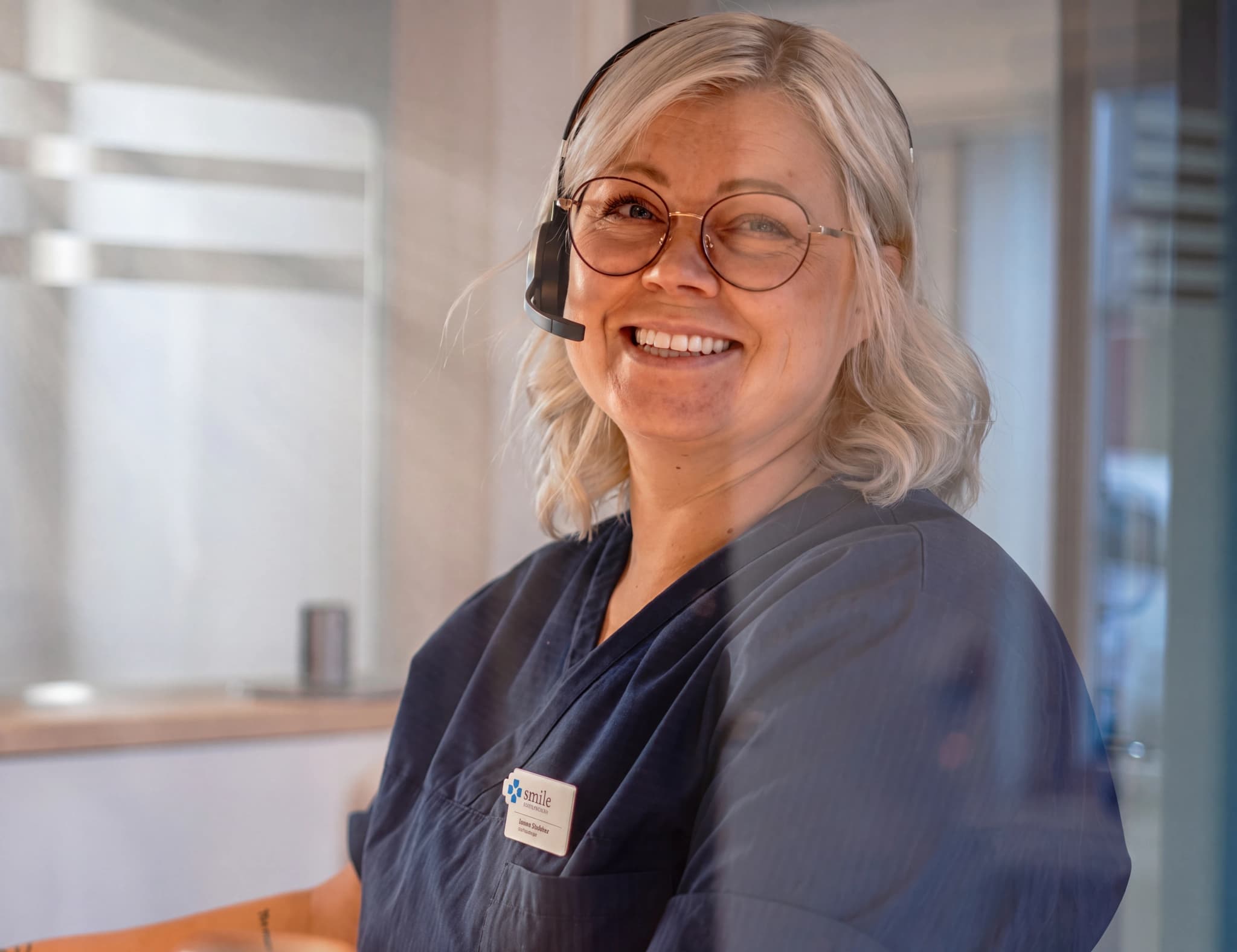 En leende kvinna med glasögon och headset står i receptionen på en klinik.