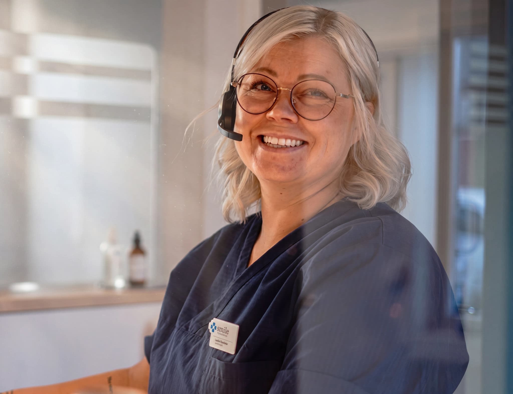 Receptionist med headset ler och tittar mot kameran på en vårdcentral.
