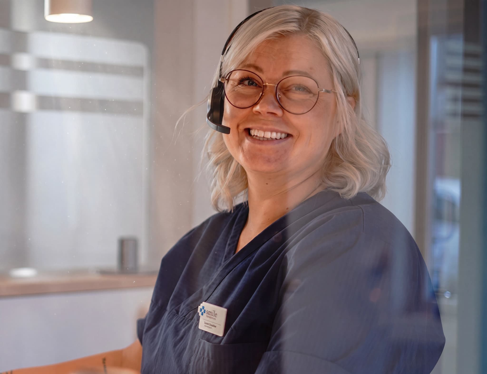 En leende kvinna med headset och glasögon bär en mörkblå klädedräkt och tittar in i kameran.