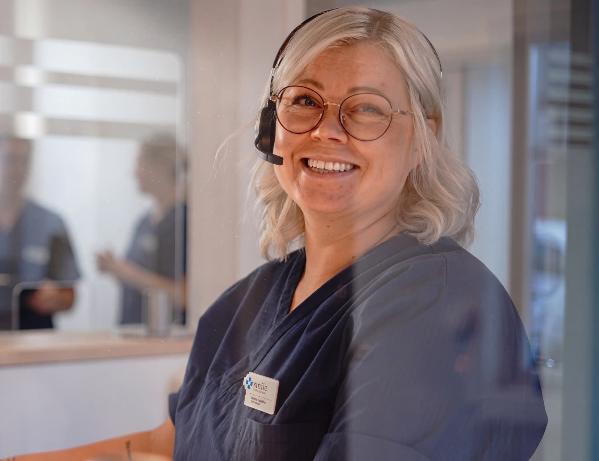 En leende kvinna med glasögon och headset bär mörkblå arbetskläder vid en reception.
