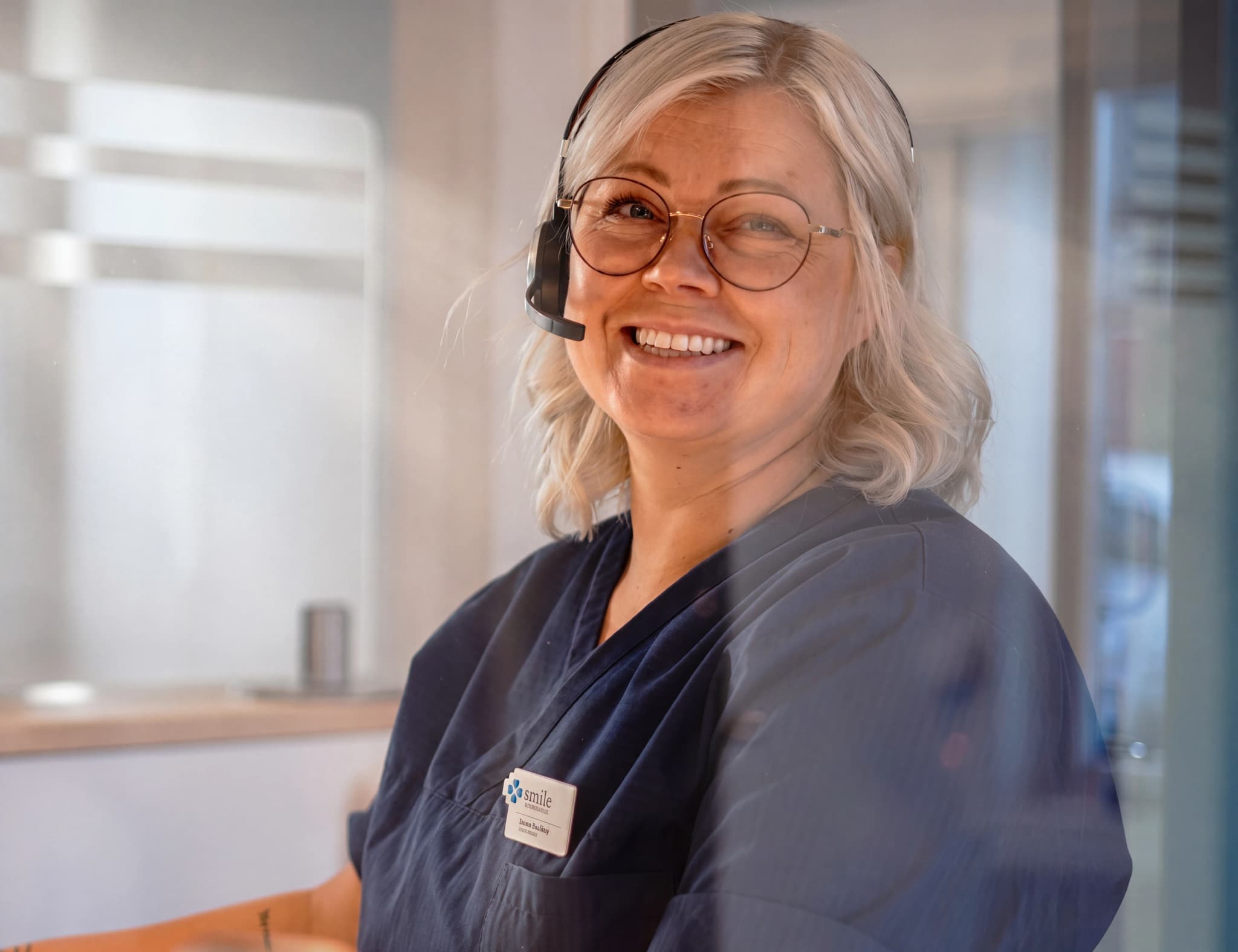 En leende receptionist med headset och glasögon bär mörkblå scrubs på en mottagning.