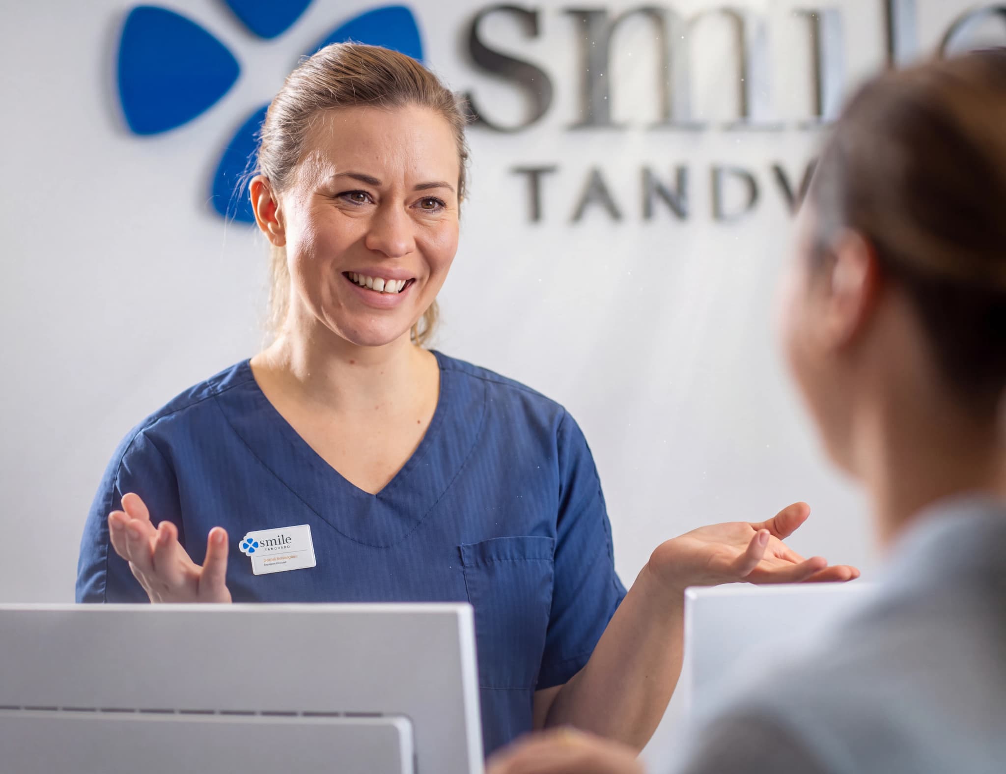 En tandläkare eller tandhygienist i blå uniform pratar med en patient vid en reception eller ett skrivbord.
