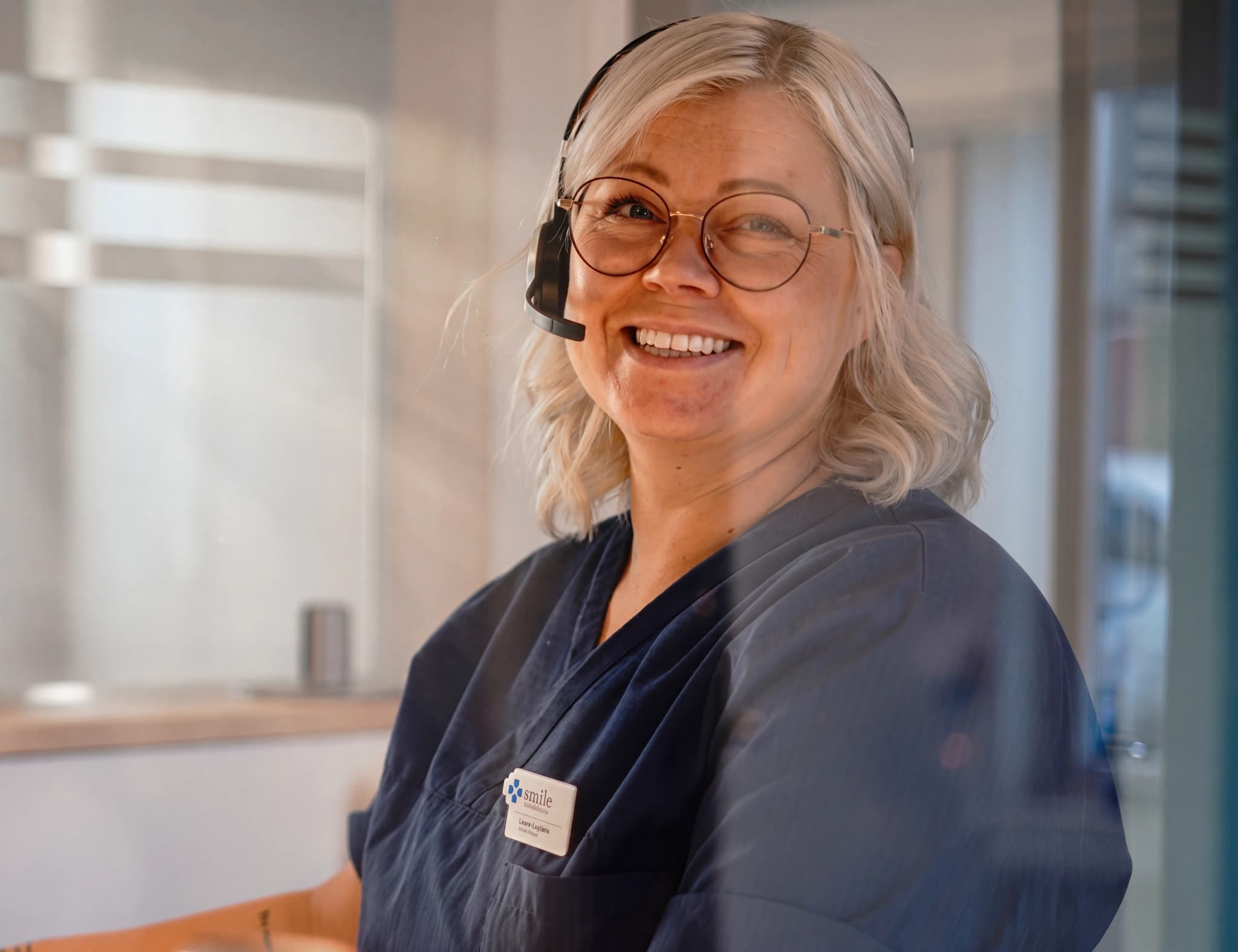 En kvinna med headset och glasögon ler mot kameran, sittande bakom en reception, förmodligen vid en medicinsk klinik.