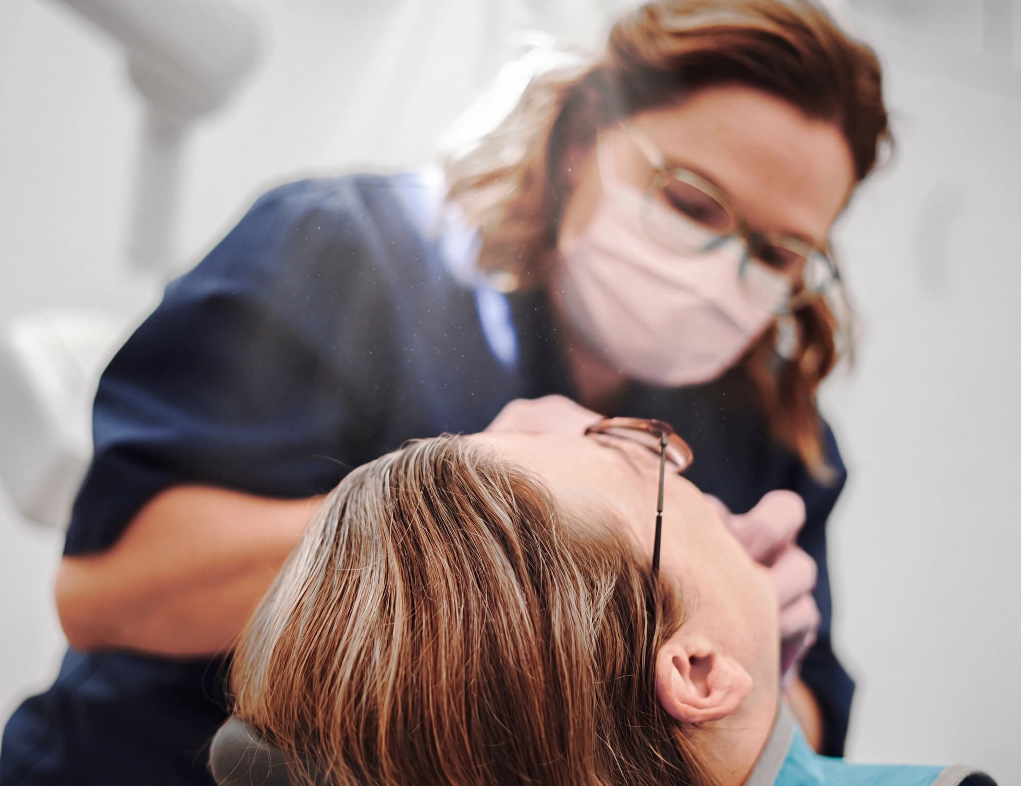 Tandläkare med munskydd och glasögon undersöker en patients tänder med ett instrument i en tandläkarstol.