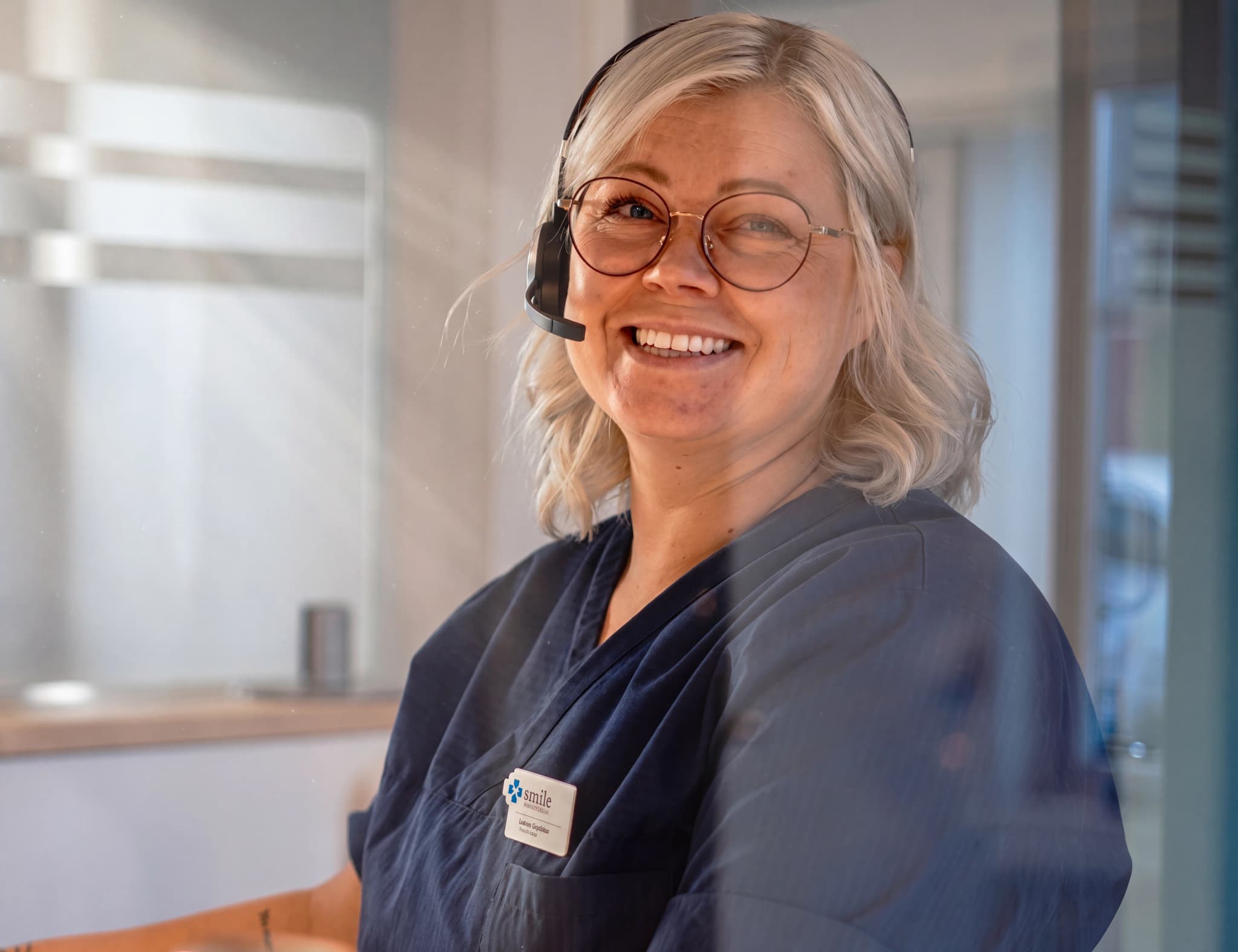 En kvinna med glasögon och headset ler mot kameran, sittandes vid ett skrivbord med en namnskylt på sin mörkblå uniform.