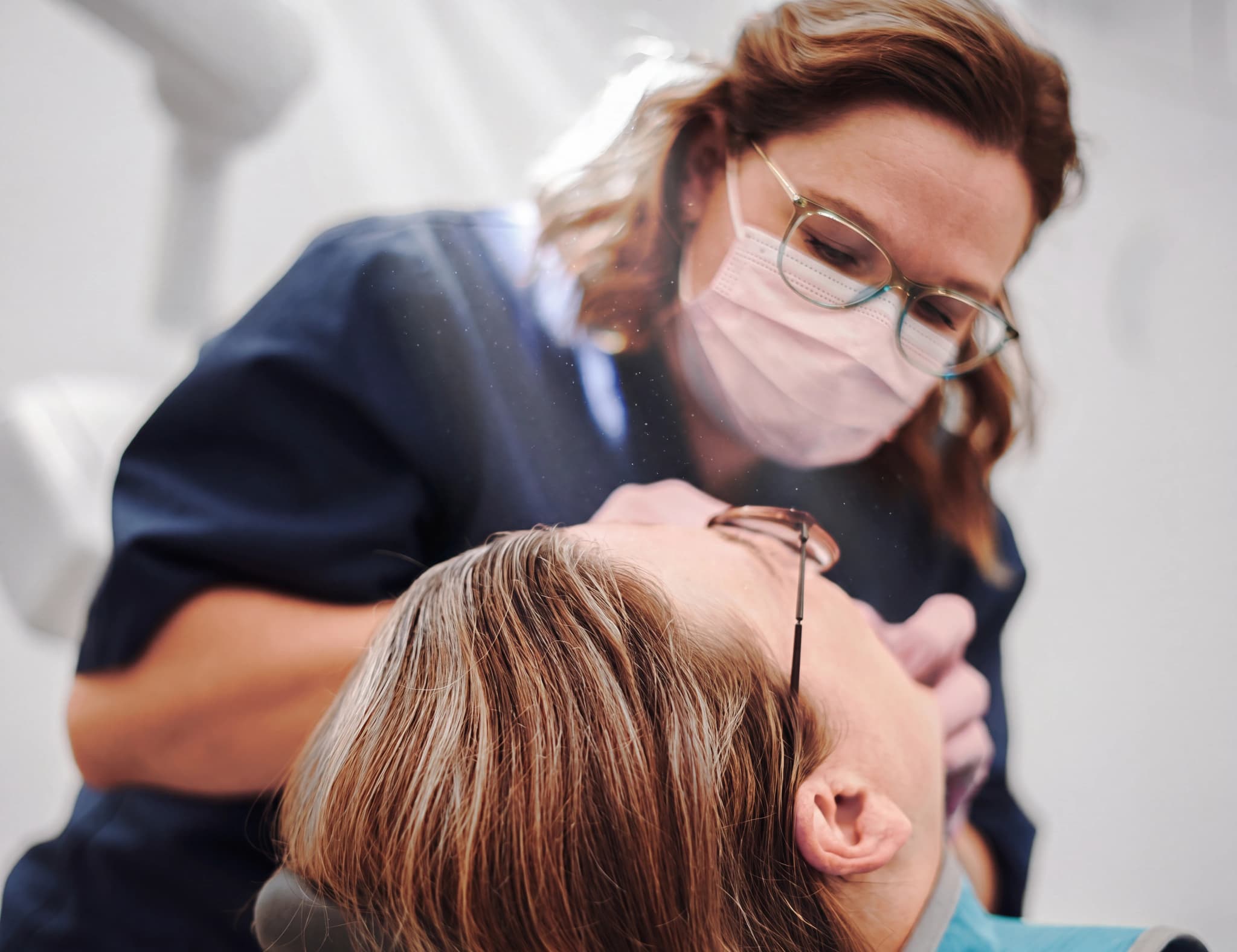 Tandläkare med mask och glasögon undersöker patientens tänder med ett instrument i en tandläkarmottagning.