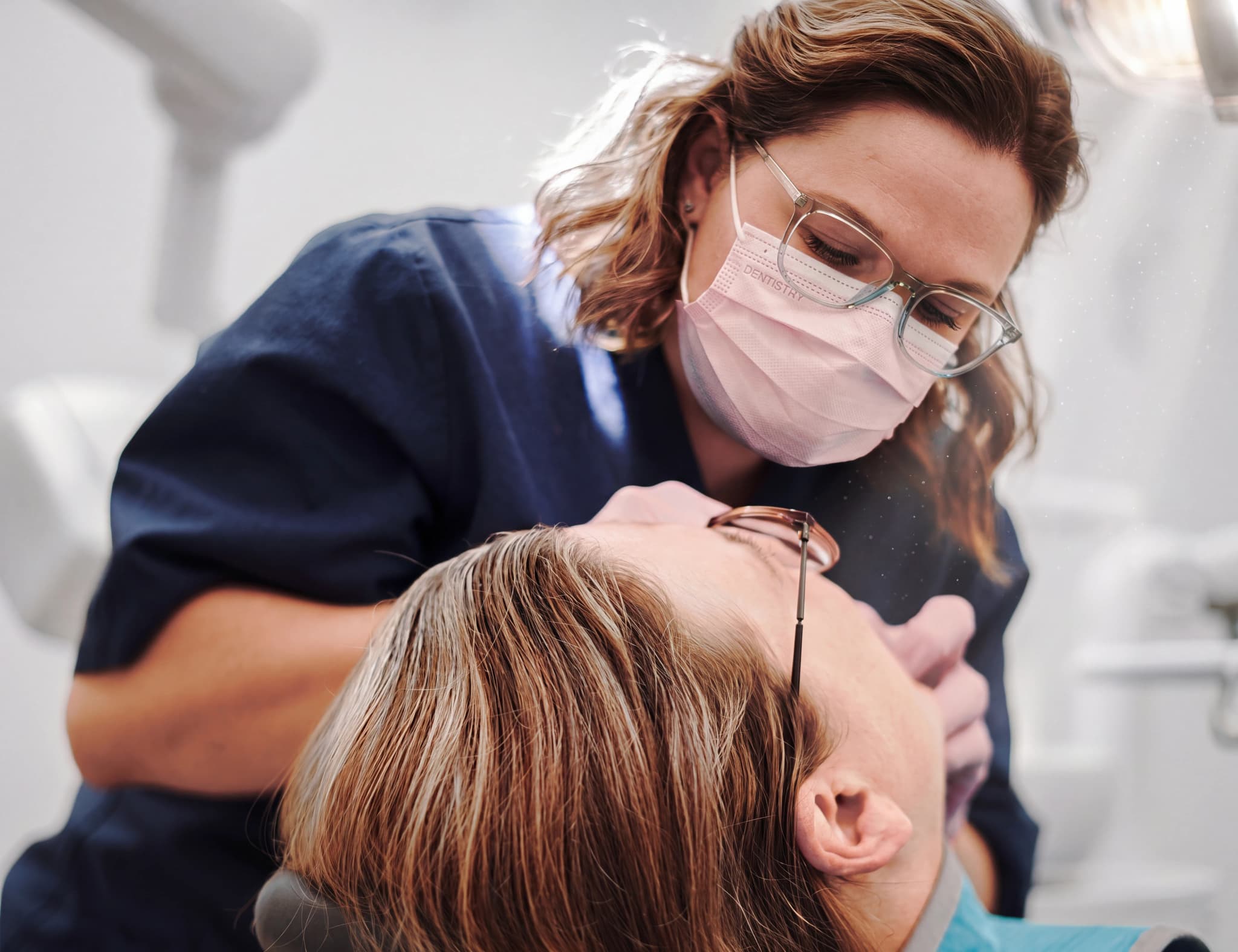 Tandläkare med munskydd och glasögon undersöker en patients tänder med ett instrument.