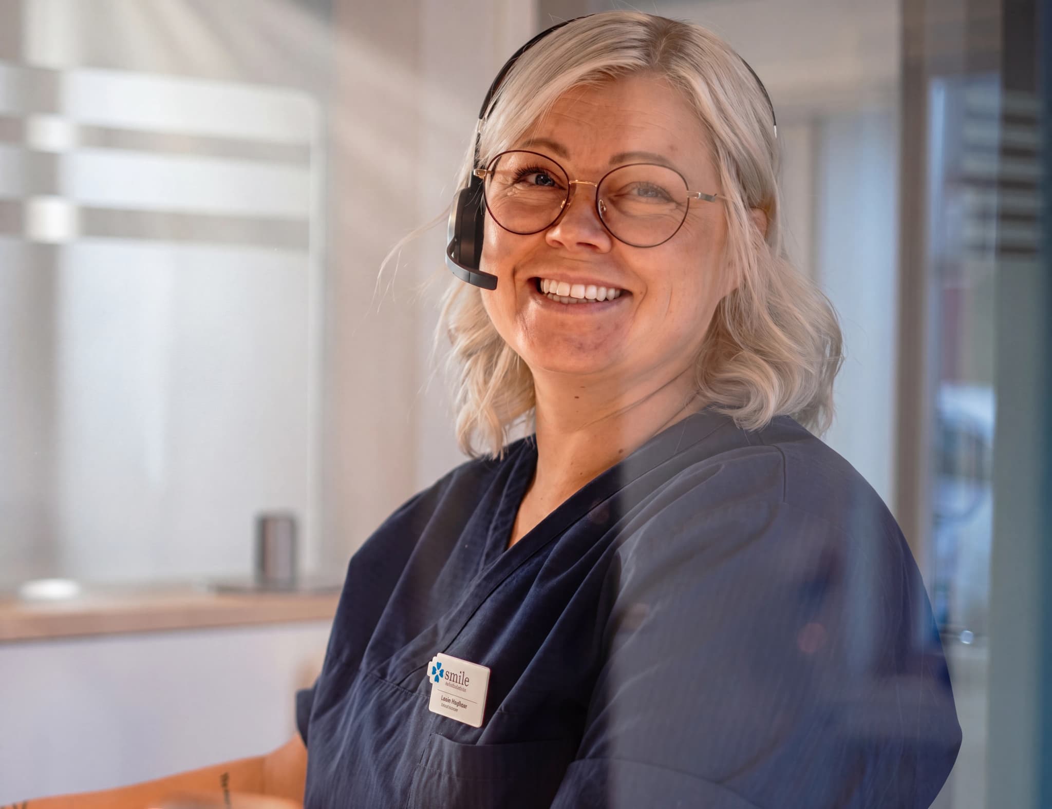 En kvinna med headset och glasögon ler mot kameran, hon är klädd i blå arbetskläder och har ett namnskylt som visar "Smile Tandvård".