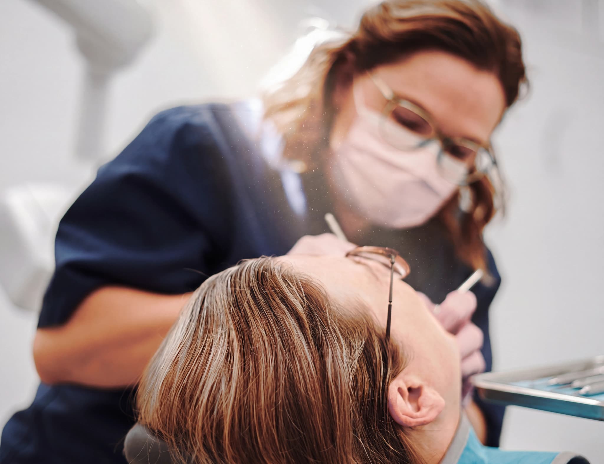 Tandläkare med munskydd och glasögon som undersöker en patients mun med ett instrument.