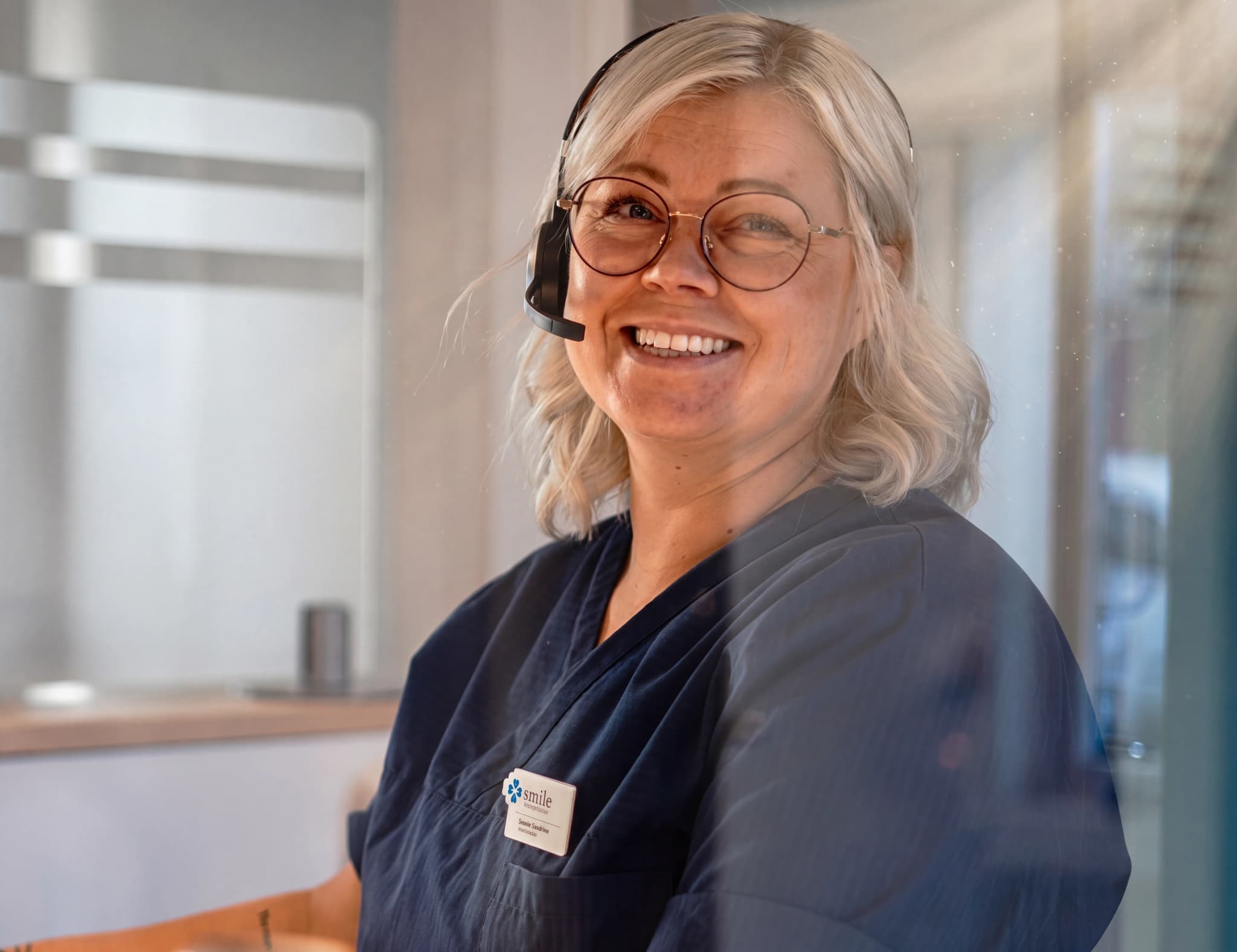 En kvinna med glasögon och headset ler mot kameran vid en reception på en klinik.