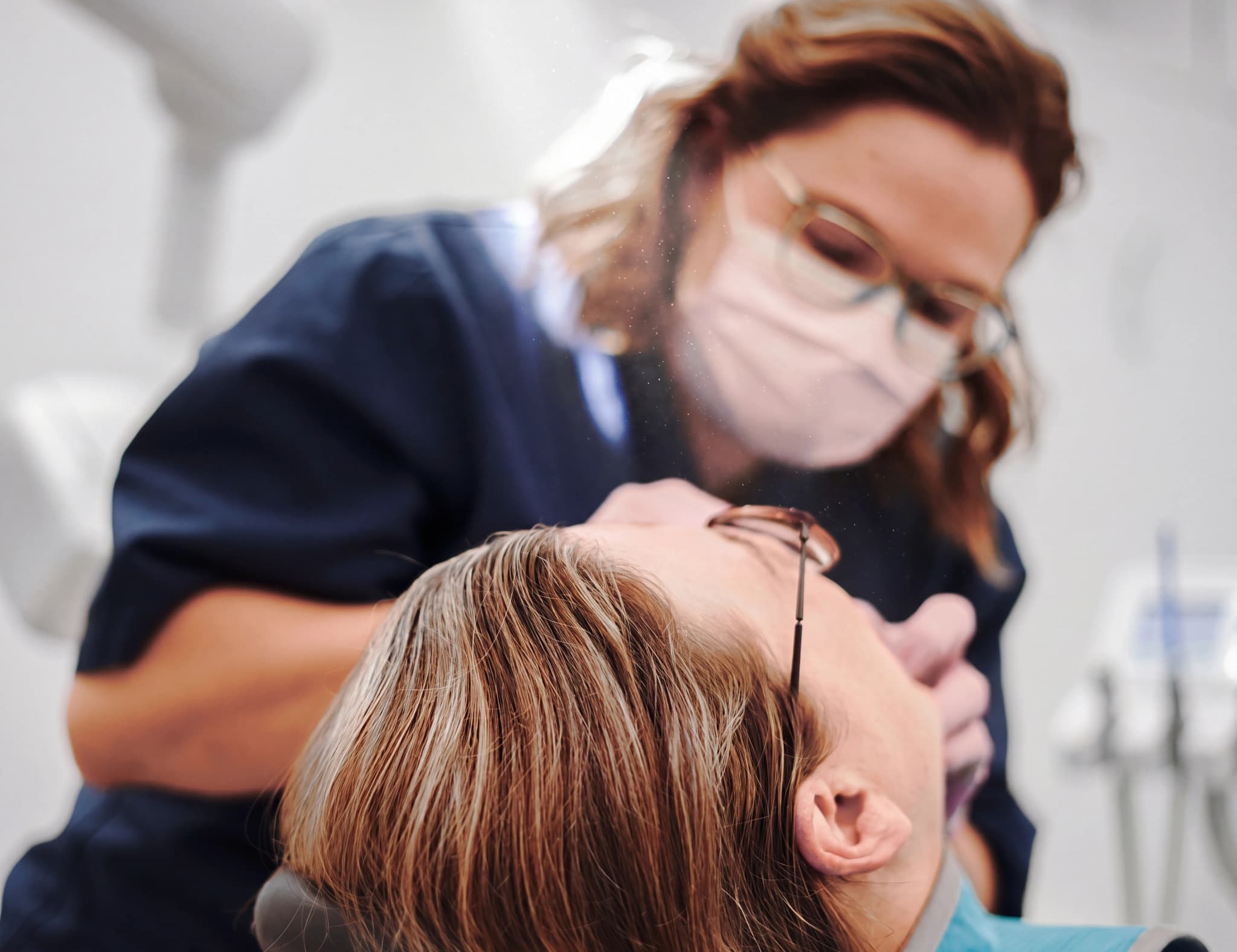 Tandläkare med mask och glasögon undersöker patientens mun med instrument i en tandläkarmottagning.