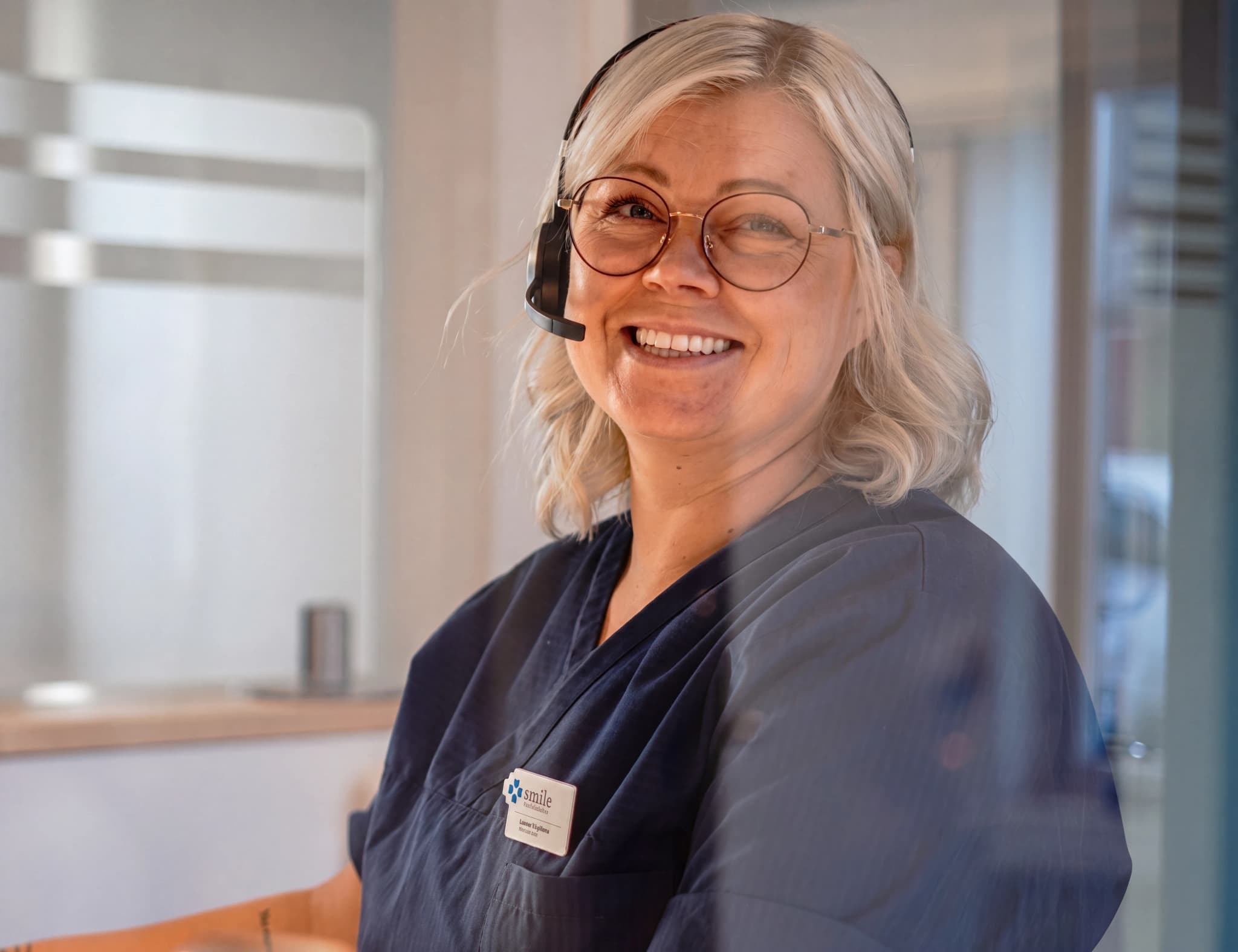 En kvinna med glasögon och headset ler mot kameran i en klinik, bärandes en blå uniform med ett namnmärke.