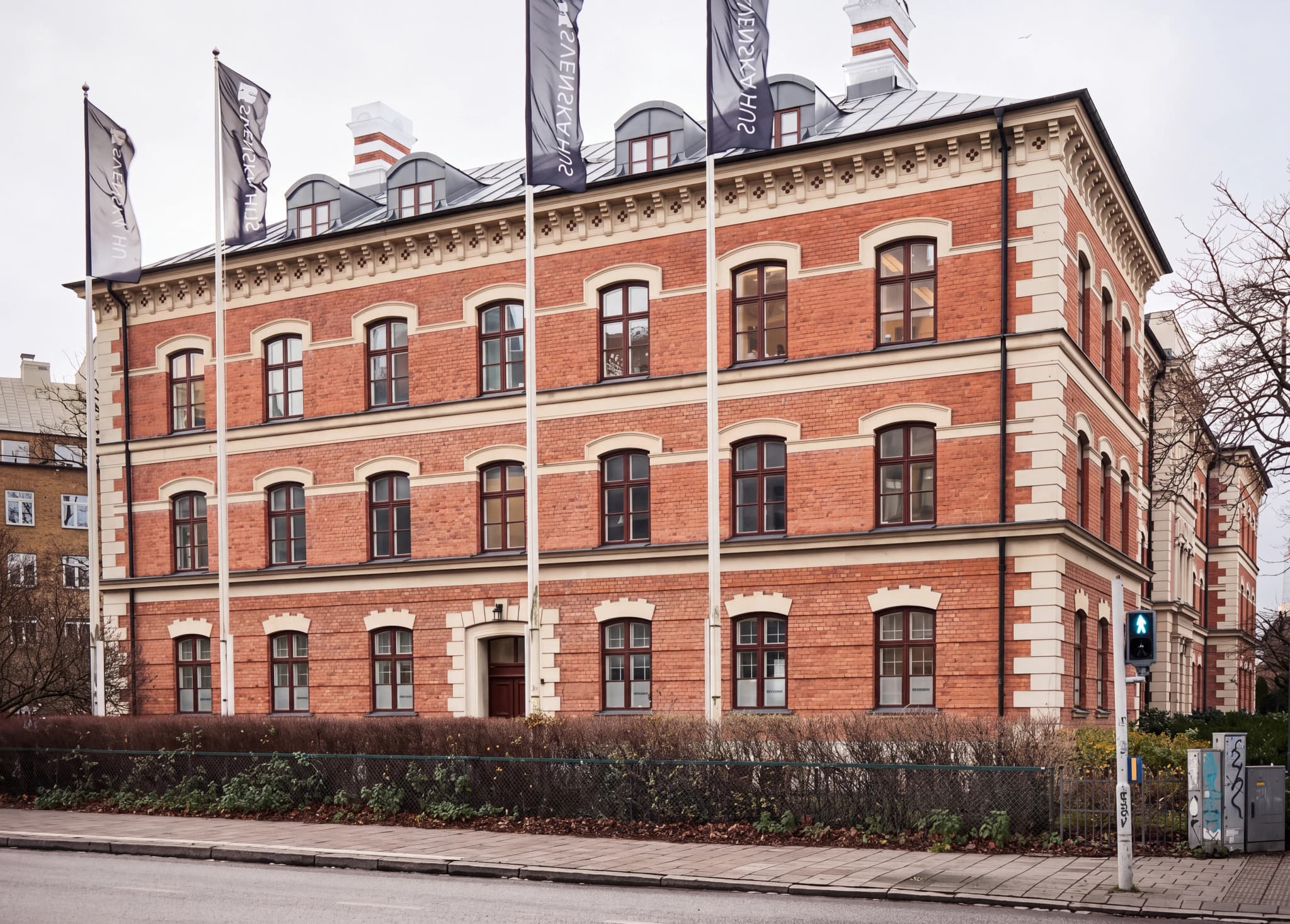 En röd tegelbyggnad med flera våningar, flaggor utanför och en trafikljus vid hörnet.