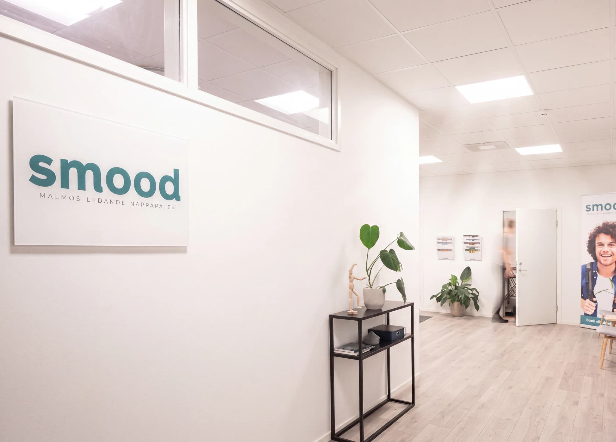 En bild på en ljus och modern klinikkorridor med en 'Smood' logotyp på väggen, växter på ett sidobord och en person som går in genom en dörr i bakgrunden.