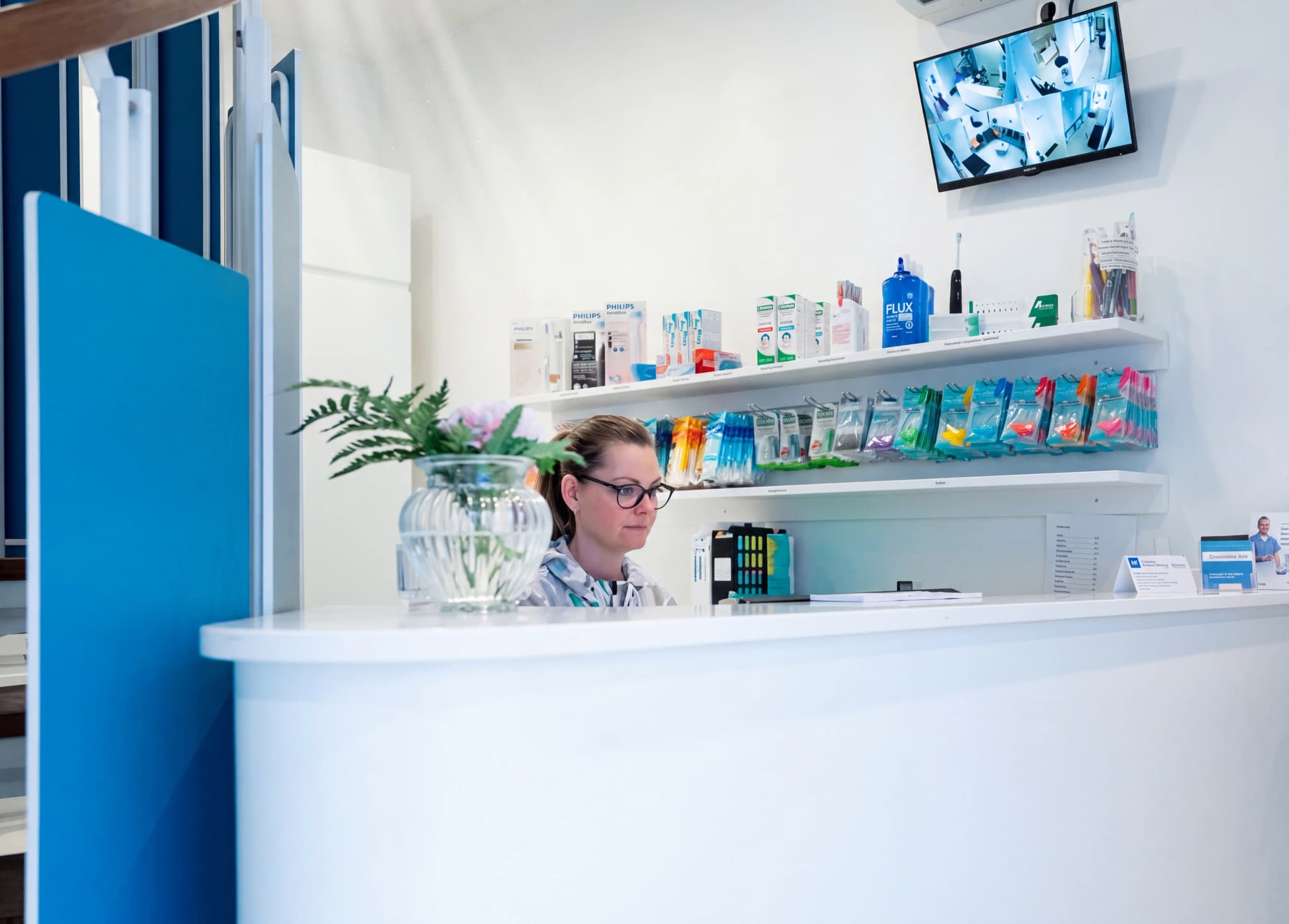 Receptionist med glasögon arbetar bakom en vit disk på en klinik, med hyllor fulla av produkter och en övervakningsskärm i bakgrunden.