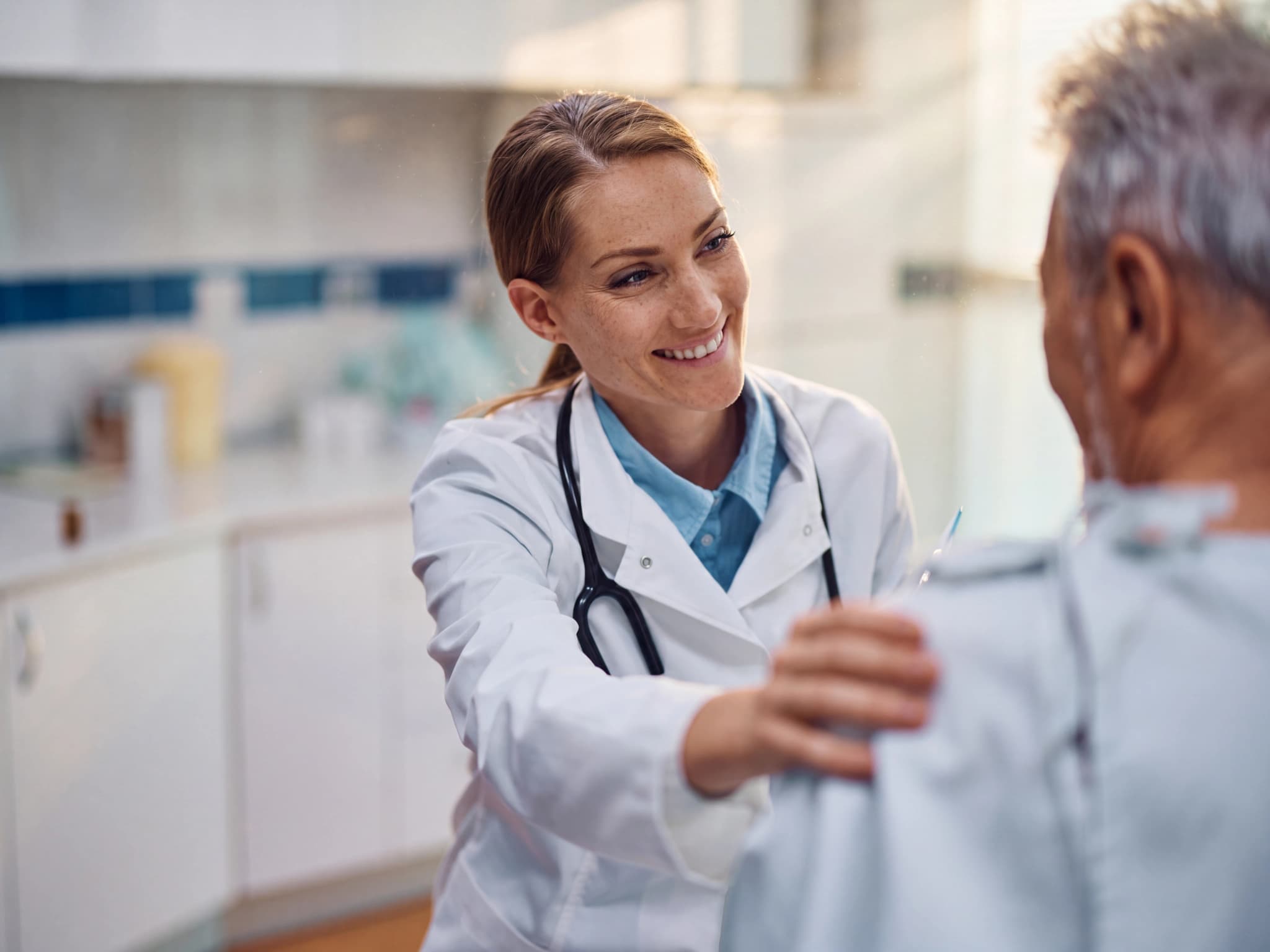Läkare ler och lägger en hand på axeln hos en äldre manlig patient under en konsultation på en klinik.
