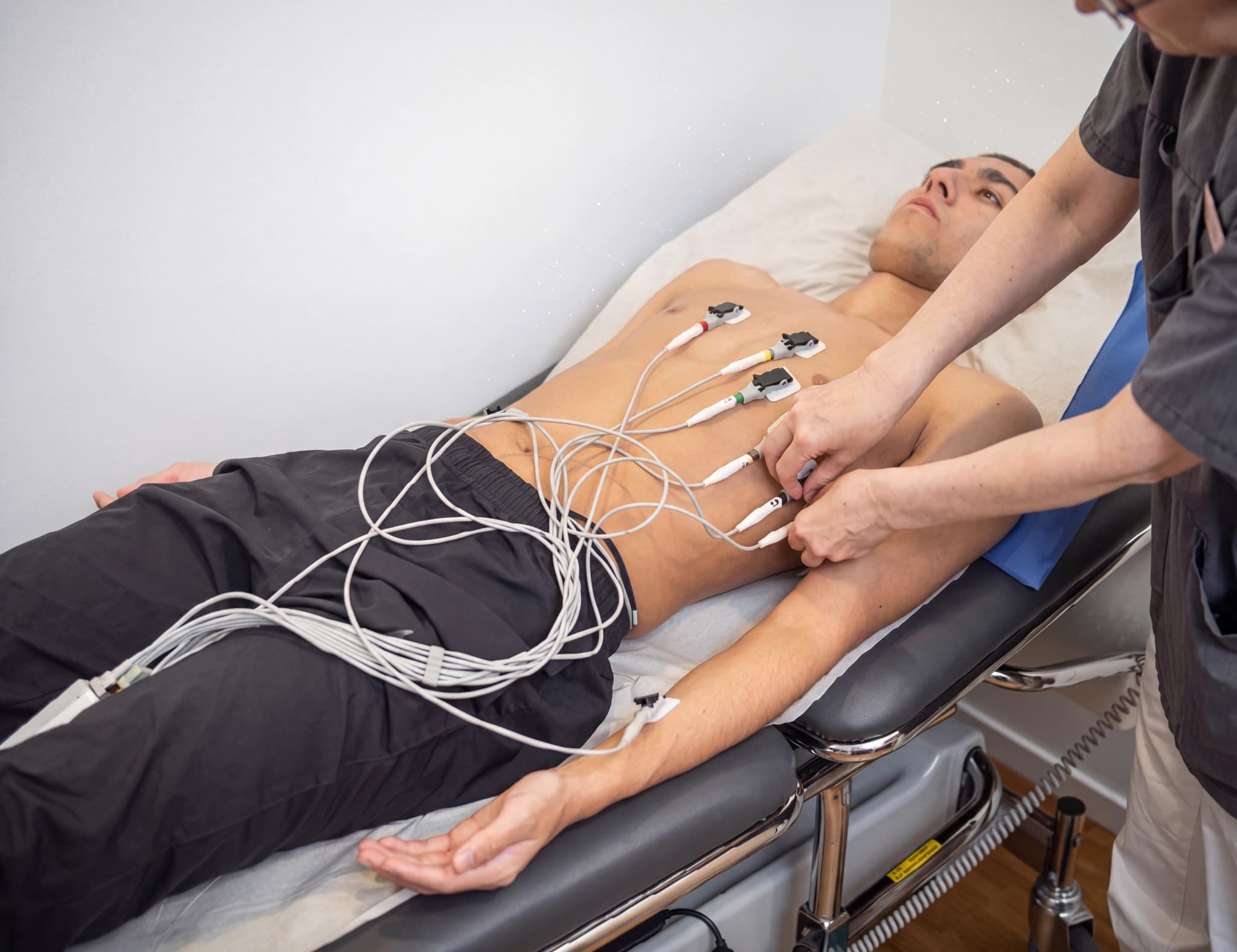 En patient ligger på en brits med elektrodsladdar fästa vid bröstet och armen medan en person i arbetskläder justerar kablarna.