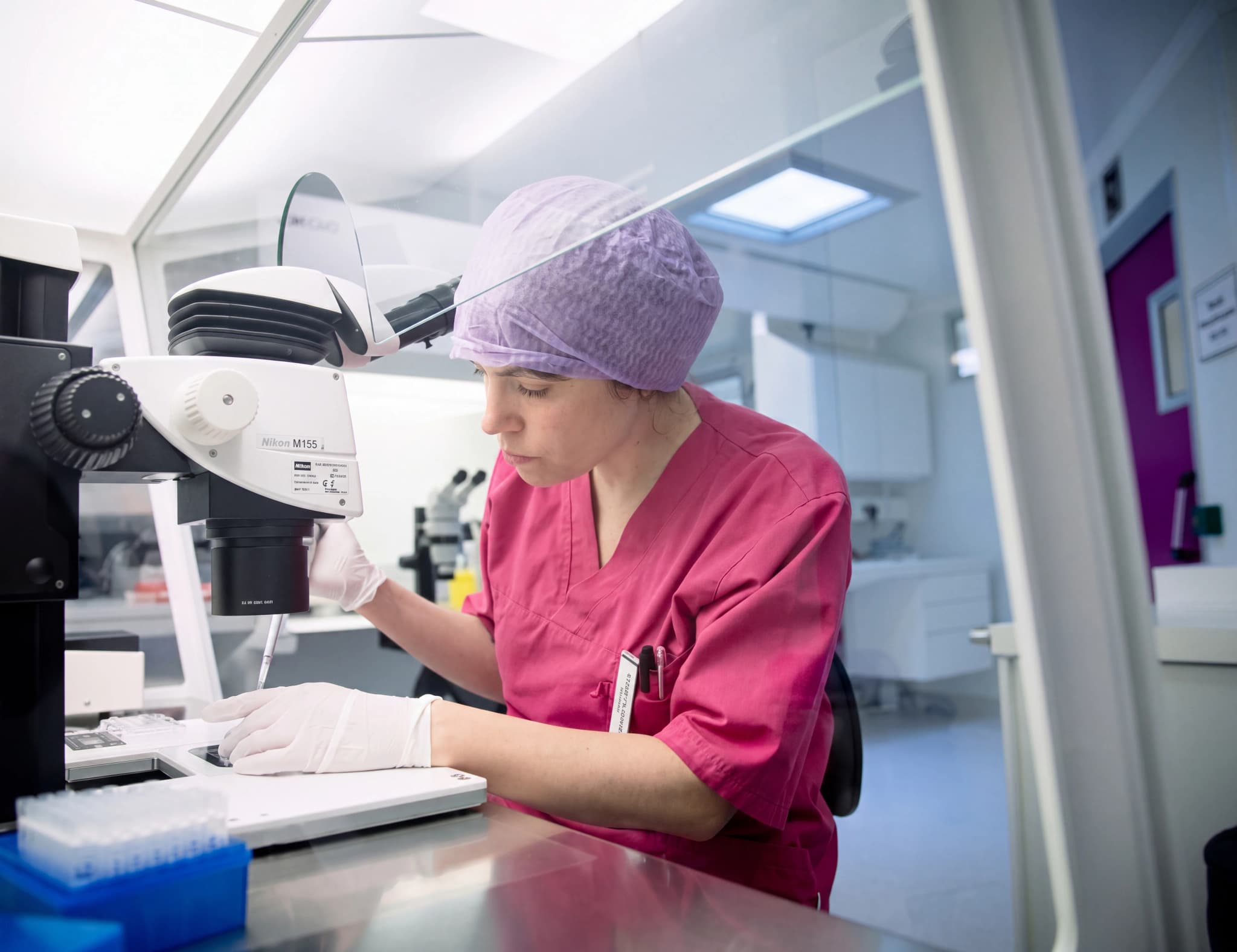 En forskare i rosa kläder och lila mössa arbetar vid ett mikroskop i ett modernt laboratorium.