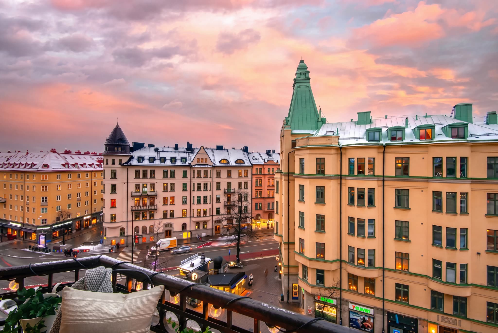 Utsikt från en balkong över en stad på vintern vid skymningen med snö på taken och upplysta fönster.