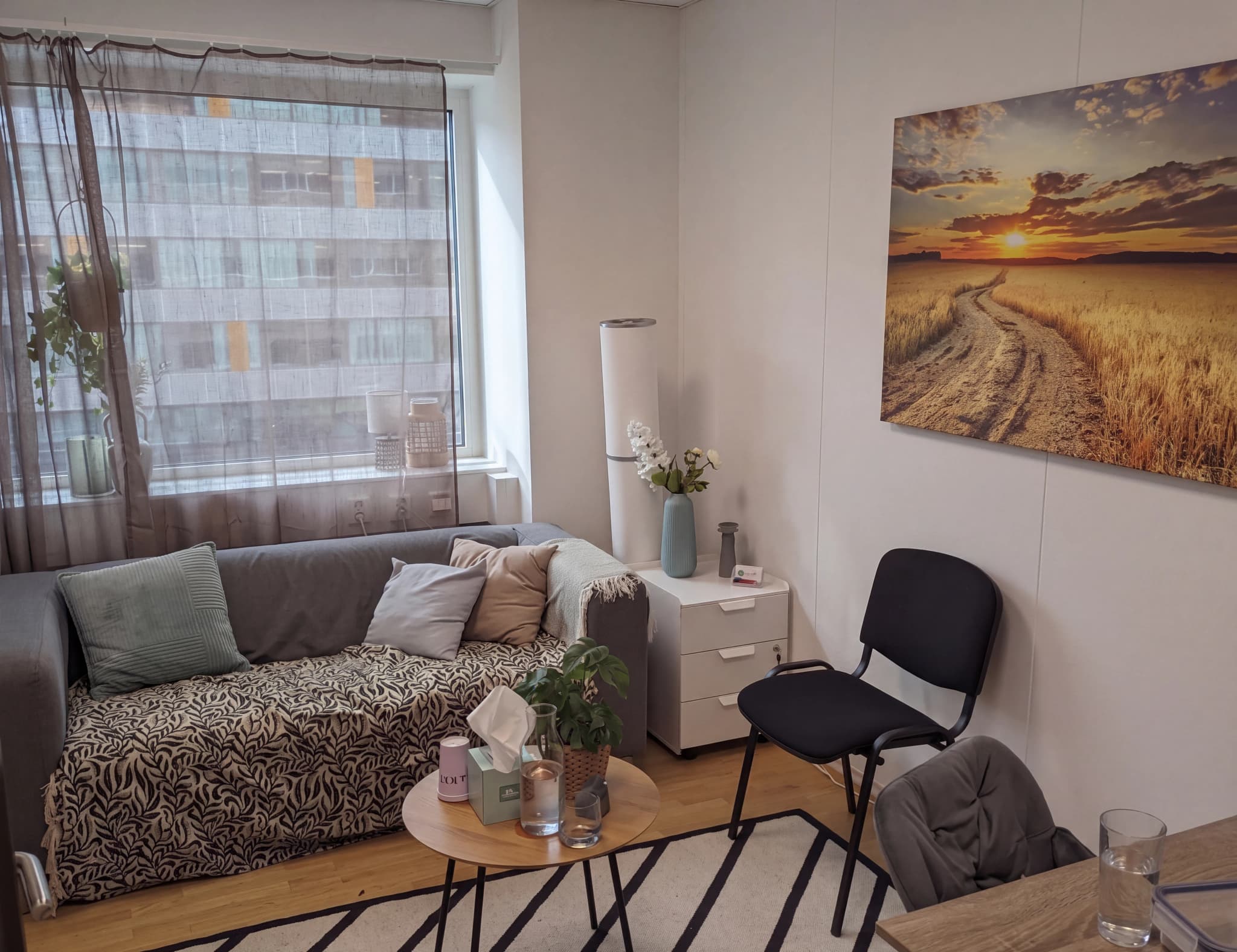 Ett terapeutiskt rum med en grå soffa, en bekväm fåtölj och väggkonst som visar en solnedgång.