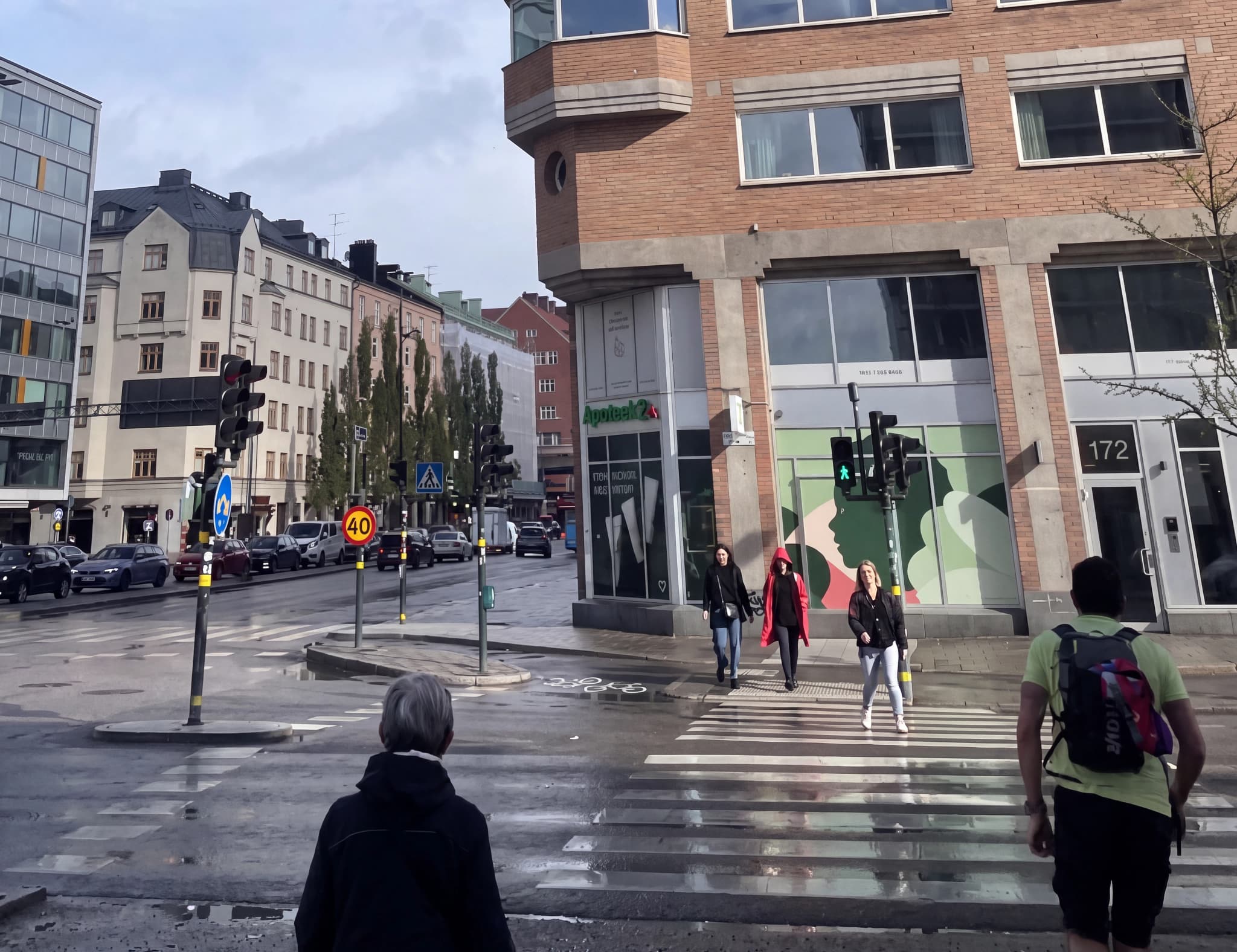 Människor korsar en gata vid en stadskorsning med trafikljus och byggnader.