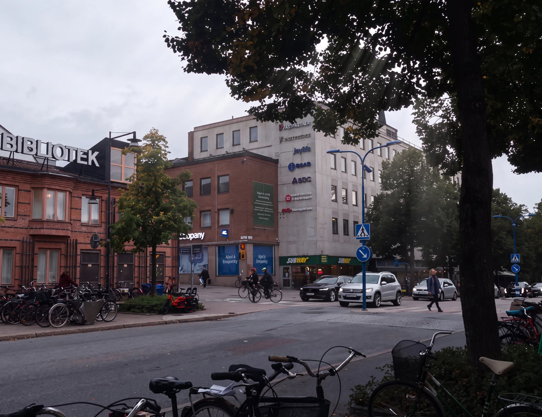 Gatubild med en byggnad som har texten BIBLIOTEK, samt andra affärsbyggnader och en gata med bilar och cyklar.