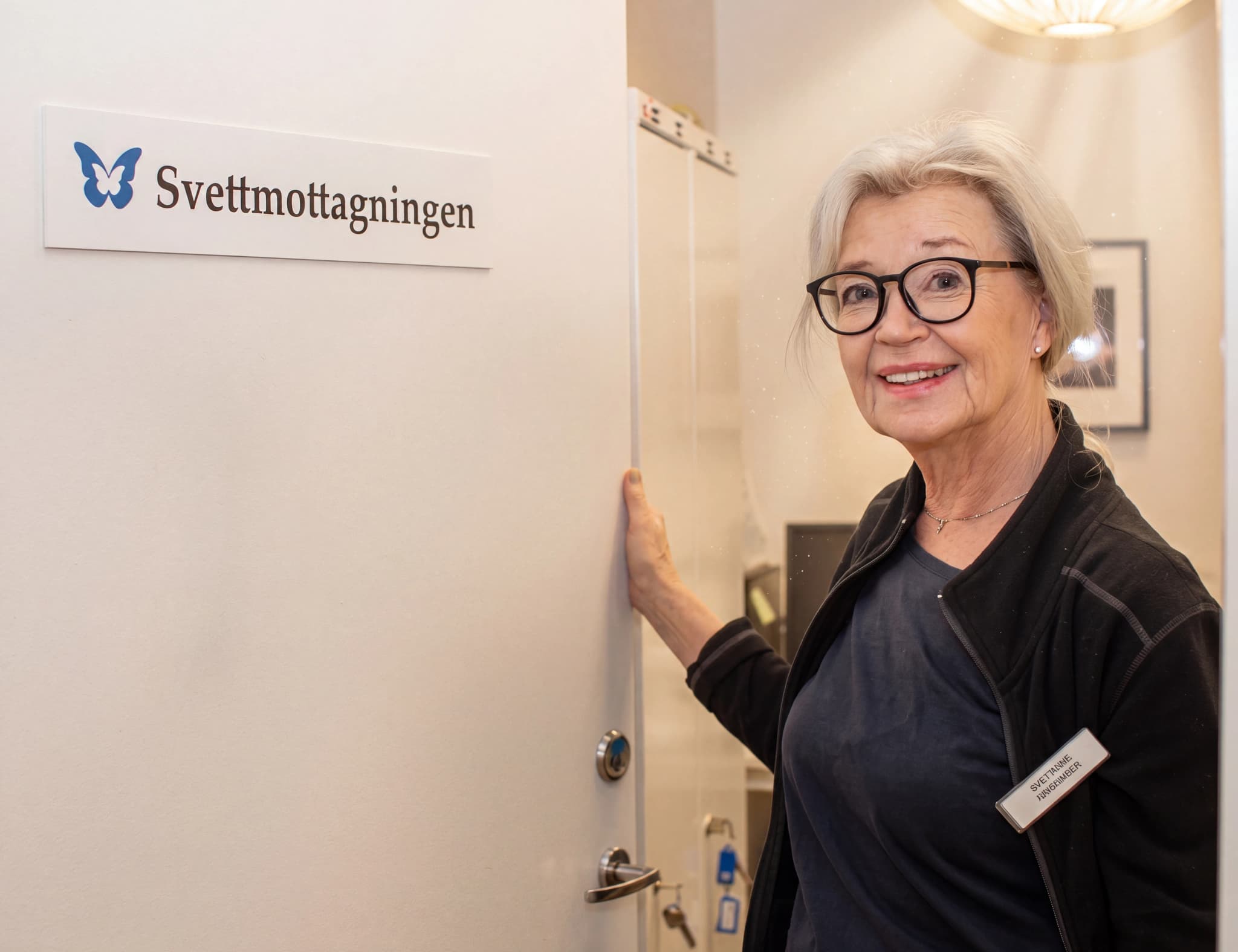 Glad kvinna i glasögon vid en dörröppning med skylten 'Svettmottagningen'.