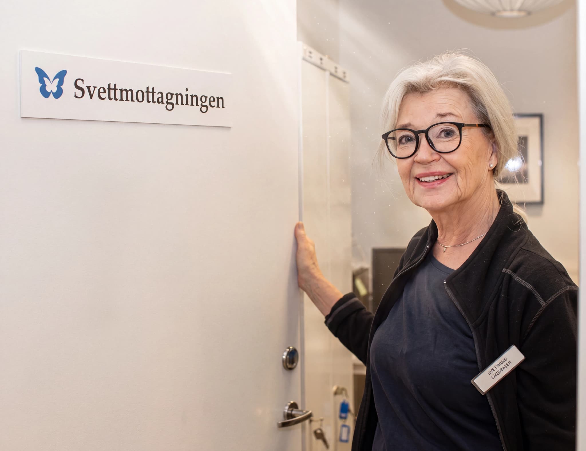 Äldre kvinna med glasögon ler och tittar in i kameran, står vid en dörröppning med en skylt som säger 'Svettmottagningen'.