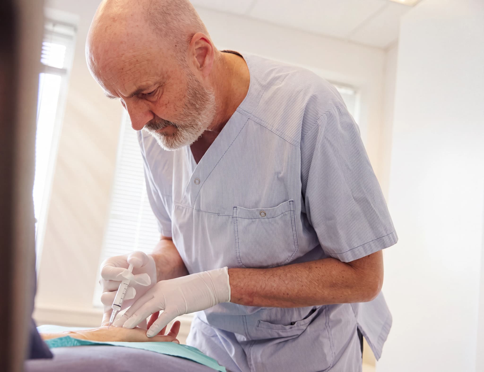 Äldre manlig läkare i ljusblå skrubbkläder ger en injektion i en patients hand med plasthandskar på, med en spruta.