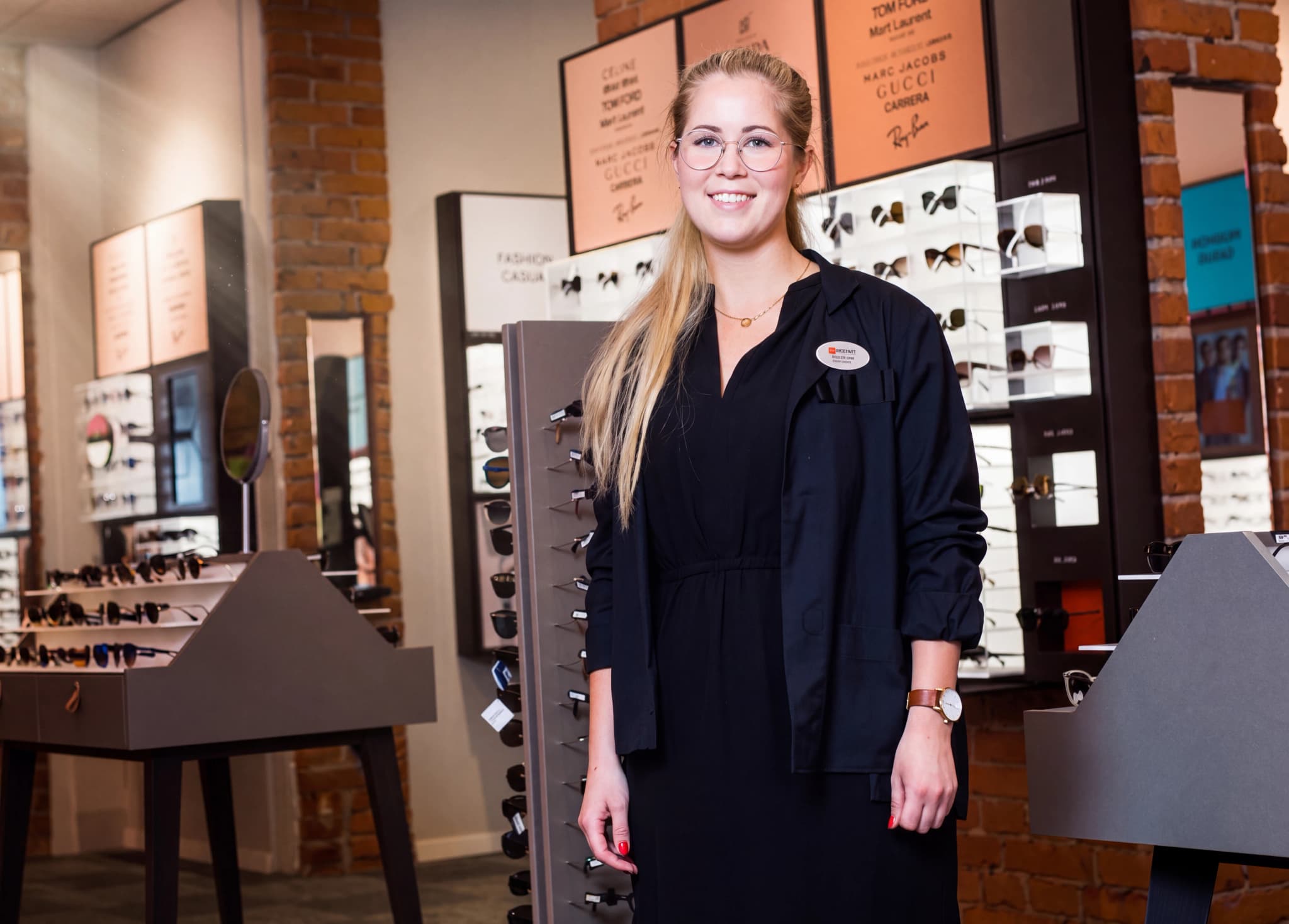 Optiker med glasögon står leende framför hyllor med glasögon i en butik.