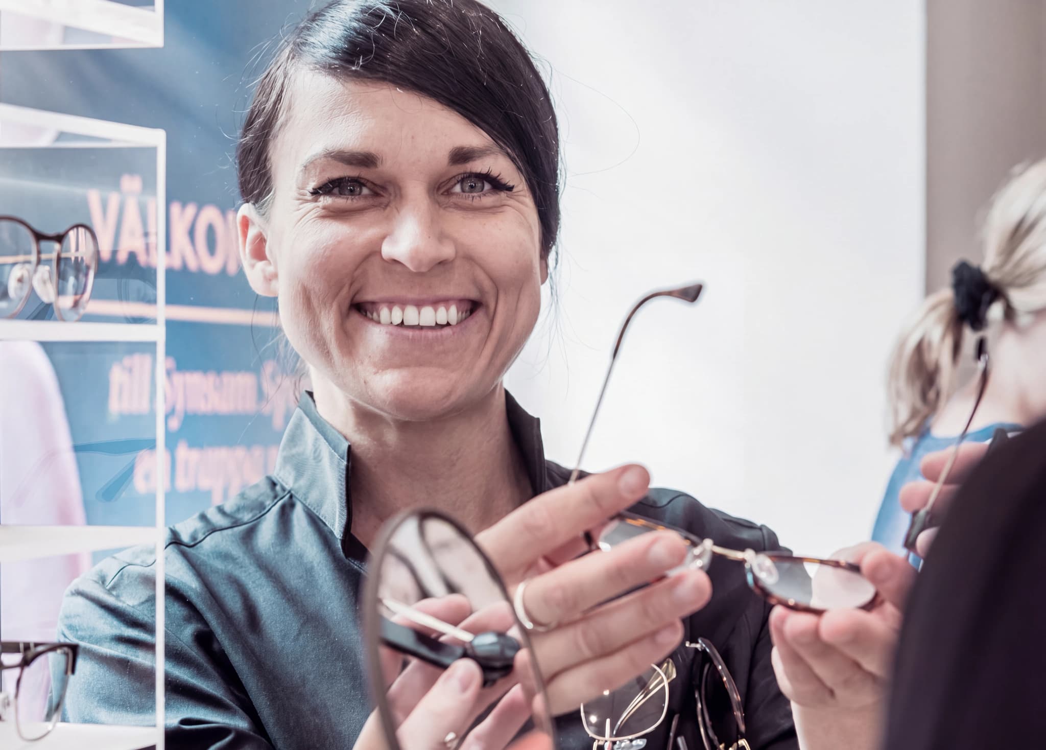 En optiker ler och håller upp ett par glasögon för en kund i en butik.