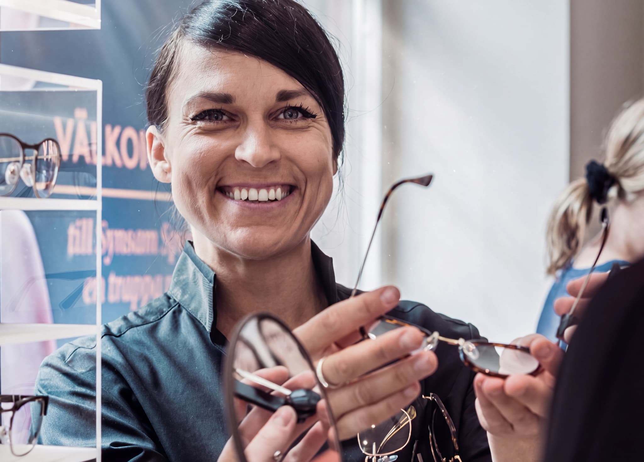 Optiker ler och visar ett par glasögon till en kund i en butik.