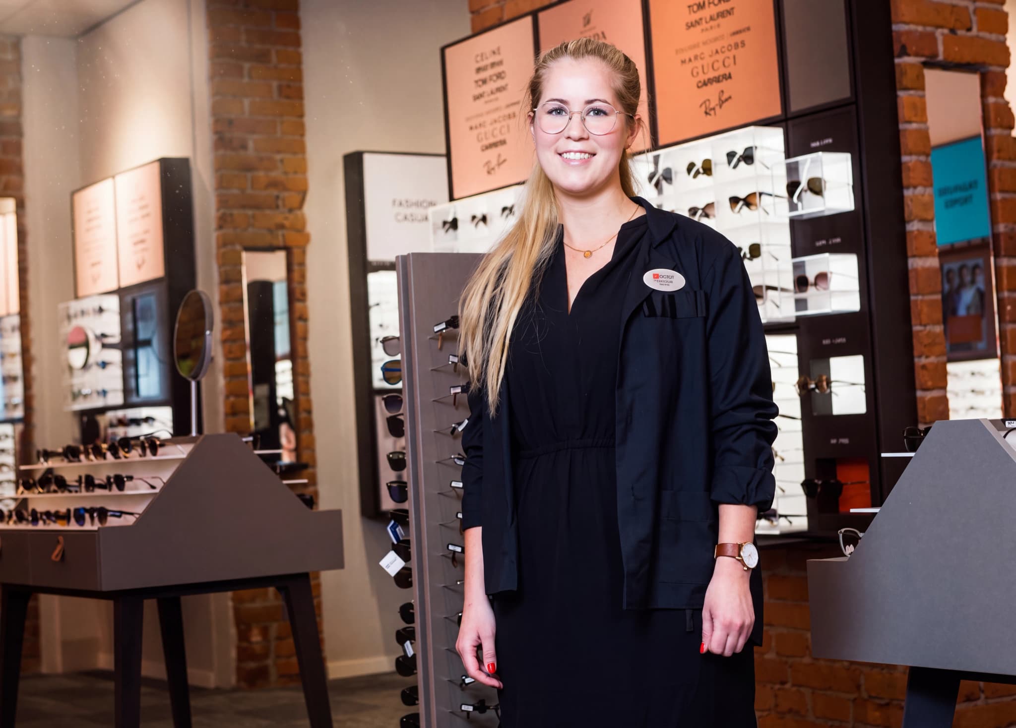 En ung kvinna med glasögon och blont hår, klädd i en svart kavaj över en klänning, står i en optikerbutik framför hyllor med glasögon.