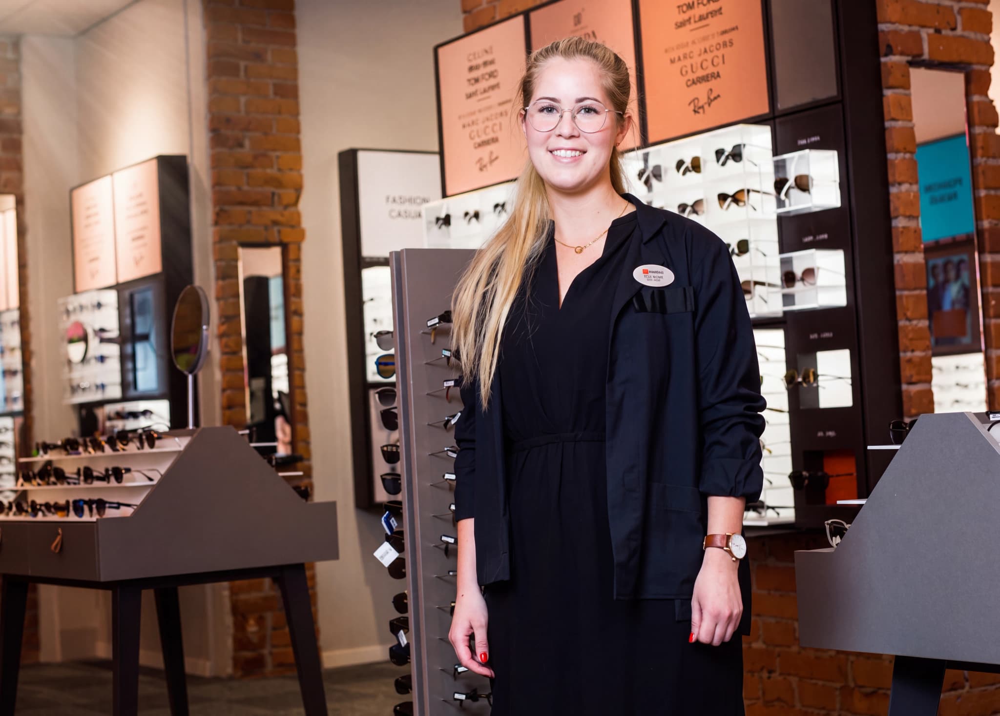 En leende ung kvinna med glasögon, klädd i mörk uniform, står i en optikaffär med hyllor fyllda med glasögon i bakgrunden.