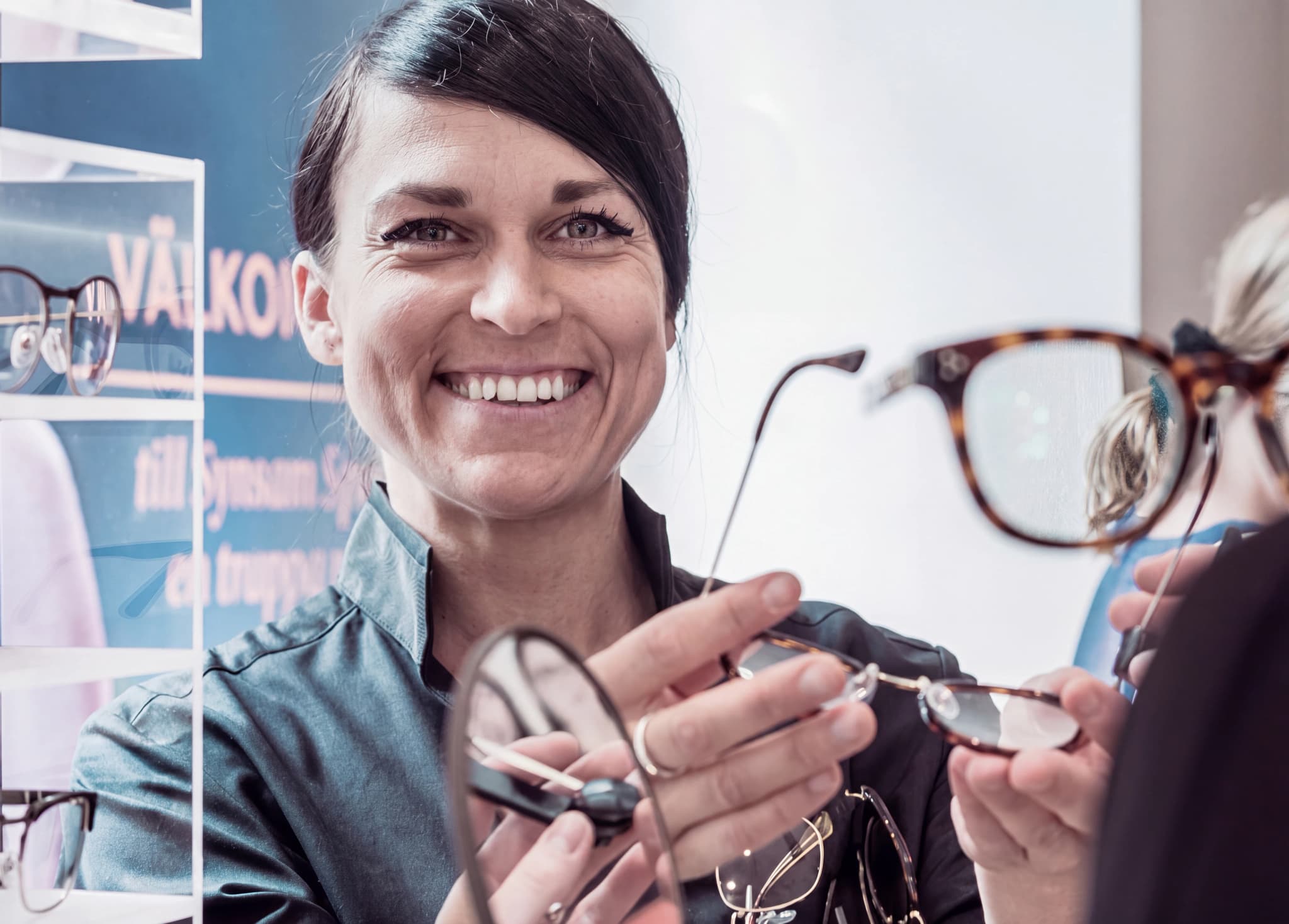 Optiker ler och visar upp ett par glasögon för en kund, med fler glasögon i bakgrunden.