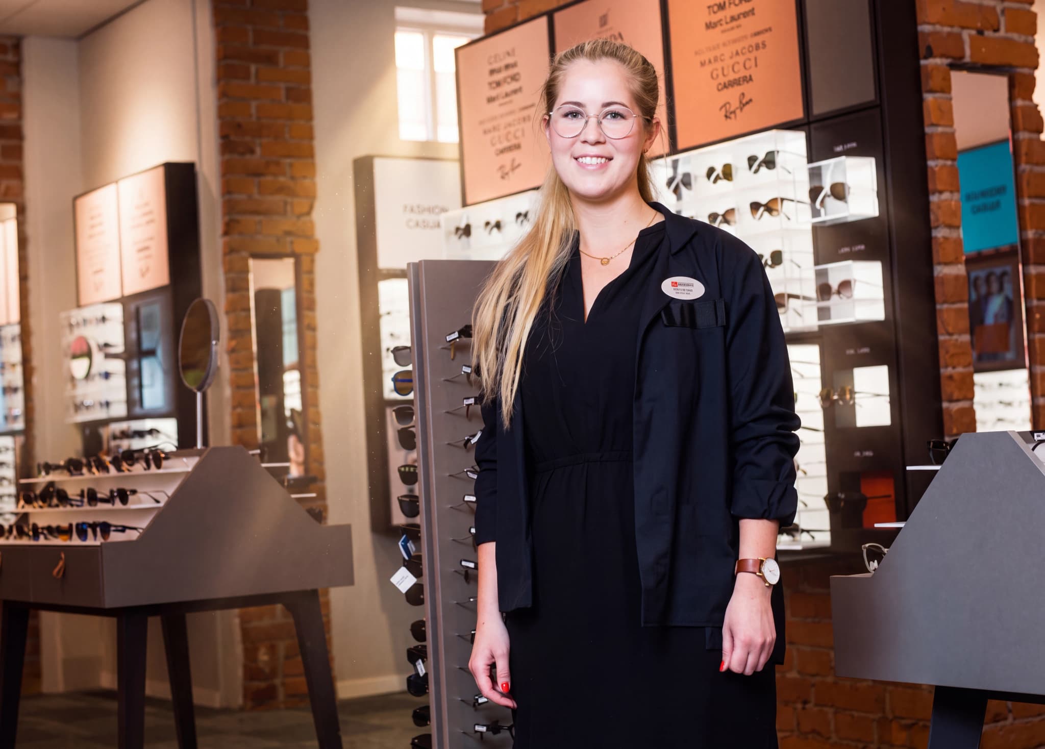 En ung kvinna med glasögon arbetar i en optikbutik och ler mot kameran.