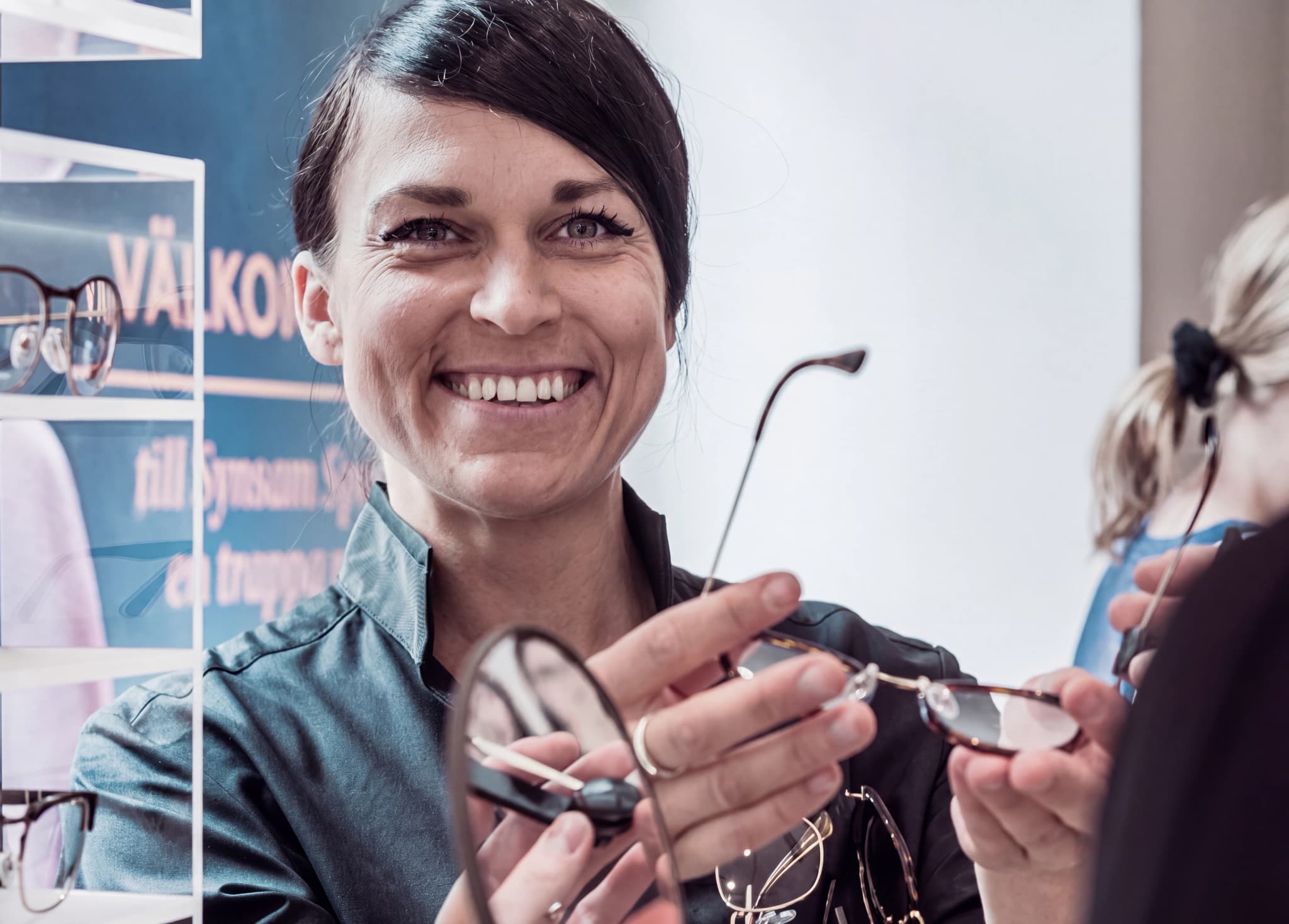 En leende optiker visar glasögon för en kund i en butik med hyllor fulla av glasögon.
