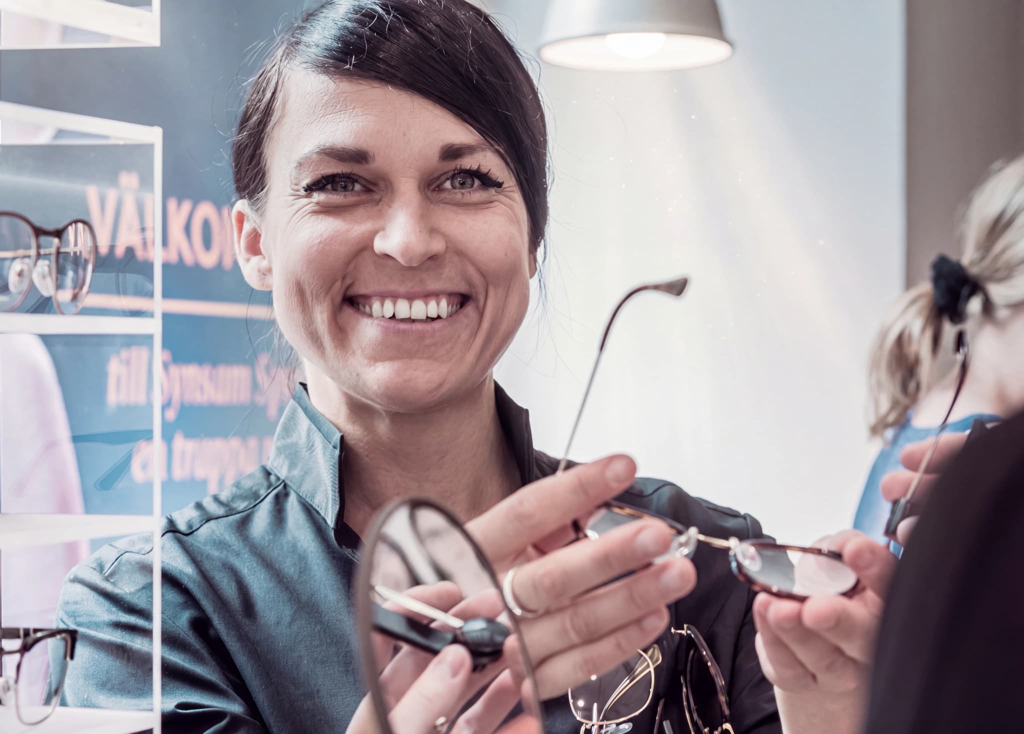 Optiker ler och visar upp ett par glasögon för en kund i en butik.