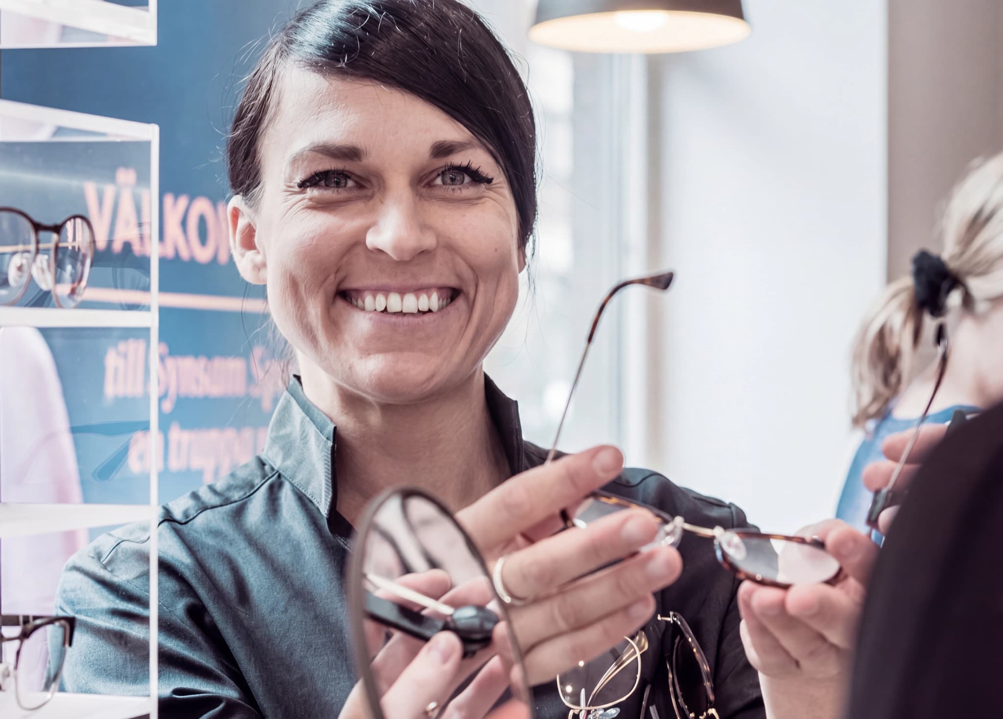 Optiker ler medan hon visar upp ett par glasögon till en kund i en butik.
