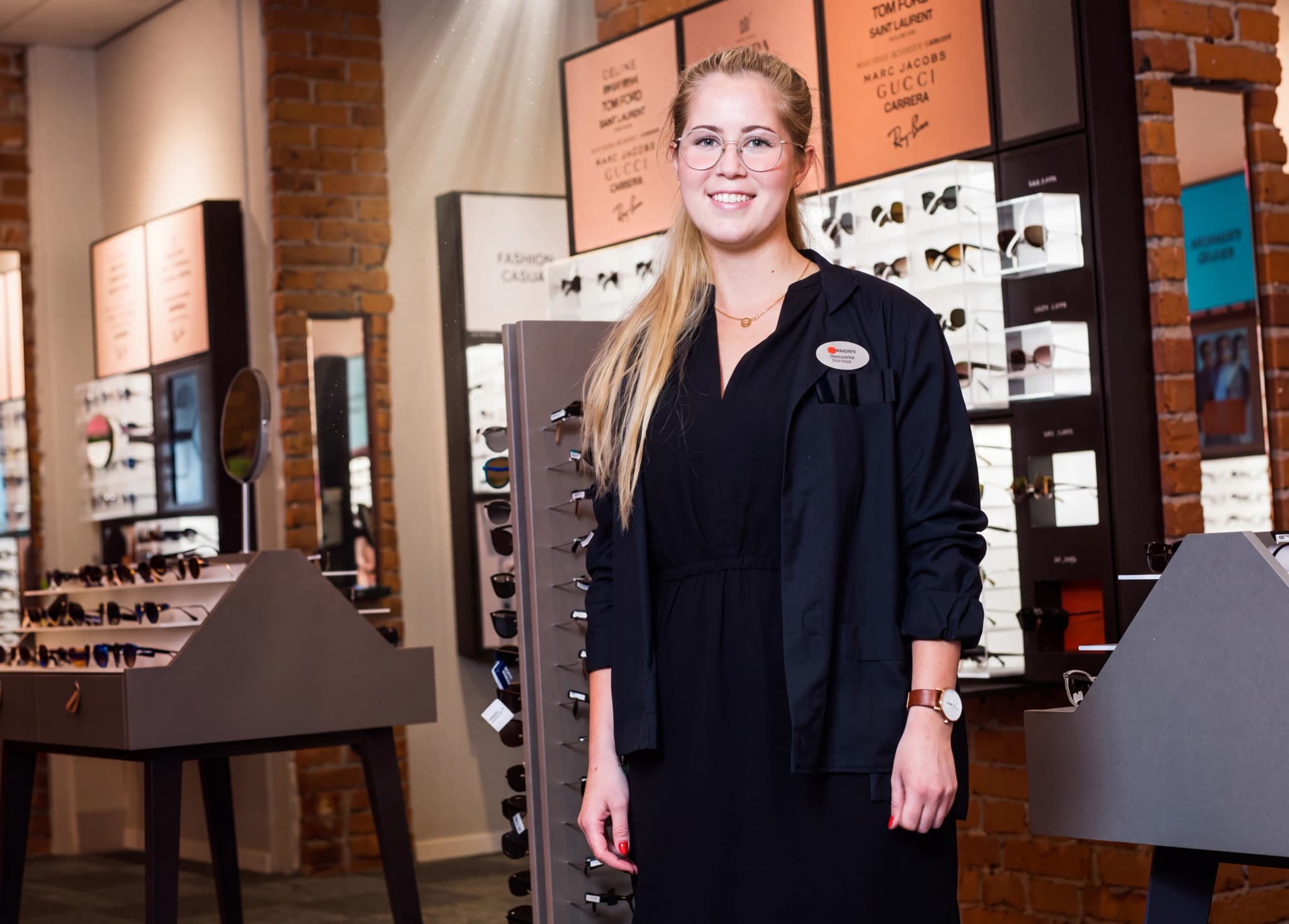En ung kvinna som arbetar i en optikerbutik står leende med glasögon framför hyllor med glasögon.