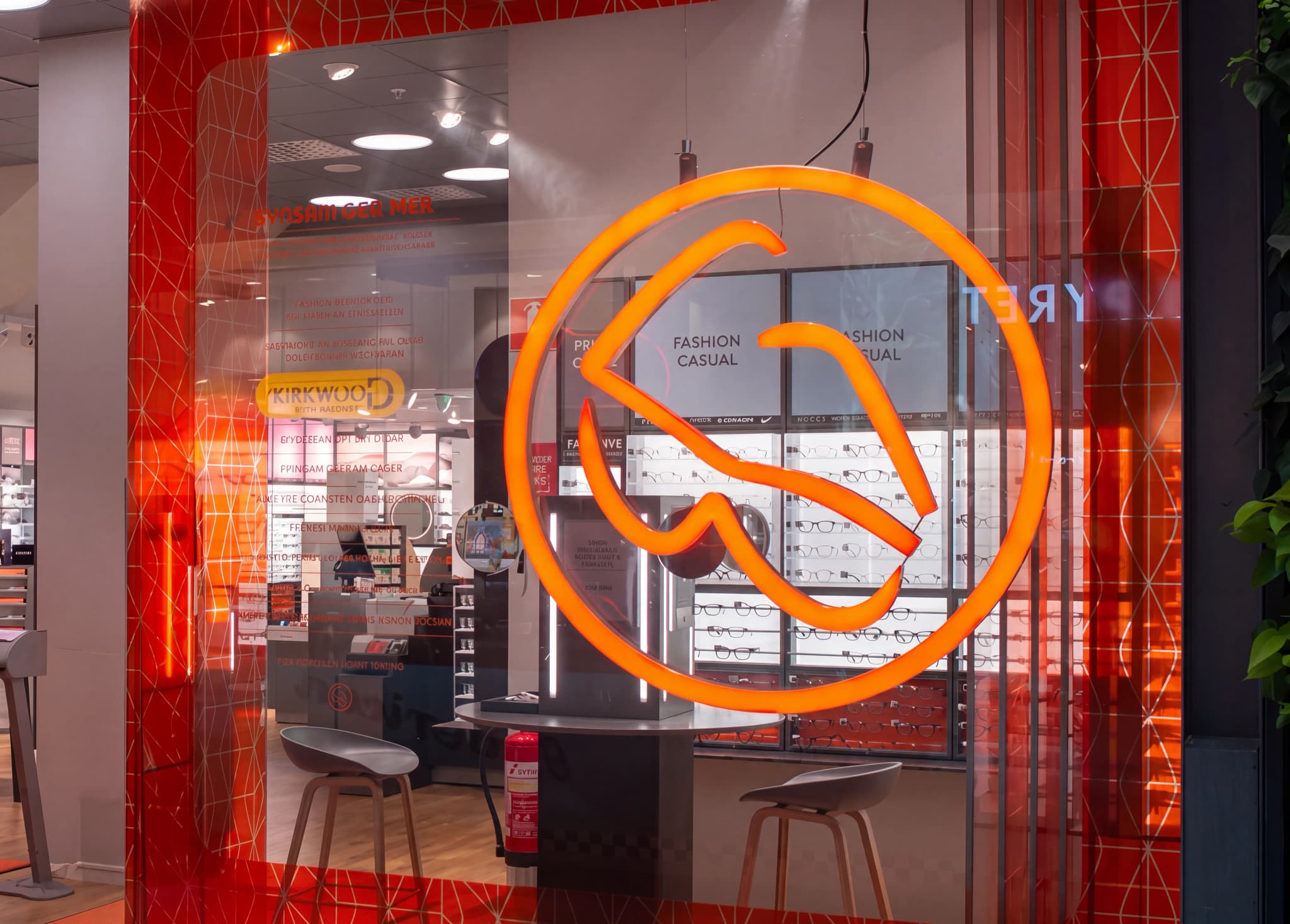 Framsidan av en optikerbutik med en stor lysande orange logotyp som visar bokstäverna 'G' och 'R', glasögon monterade bakom och två stolar framför. Fönstret har ett rött geometriskt mönster.