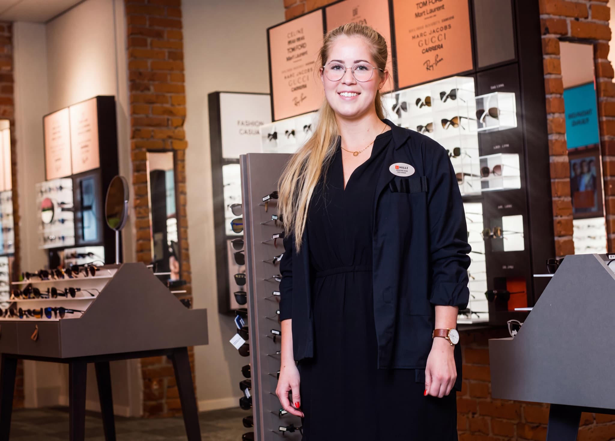 En ung kvinna med glasögon står i en butik med solglasögon, klädd i en mörk uniform.