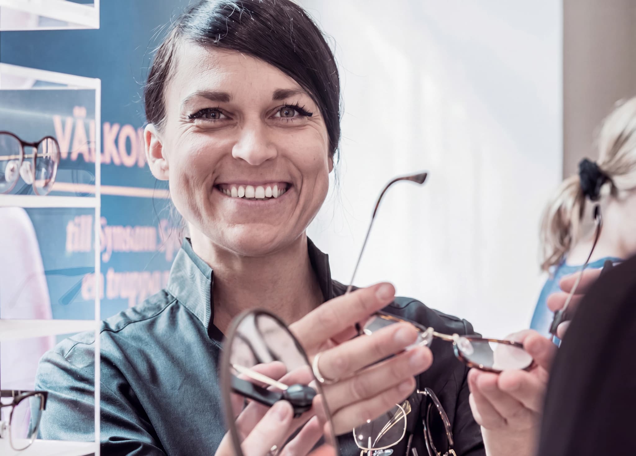 En optiker ler och visar ett par glasögon till en kund, med andra glasögon synliga i bakgrunden.