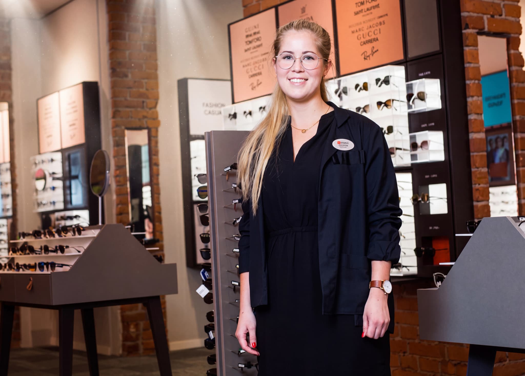 En leende optiker i mörk klädsel står framför hyllor med glasögon i en butik.