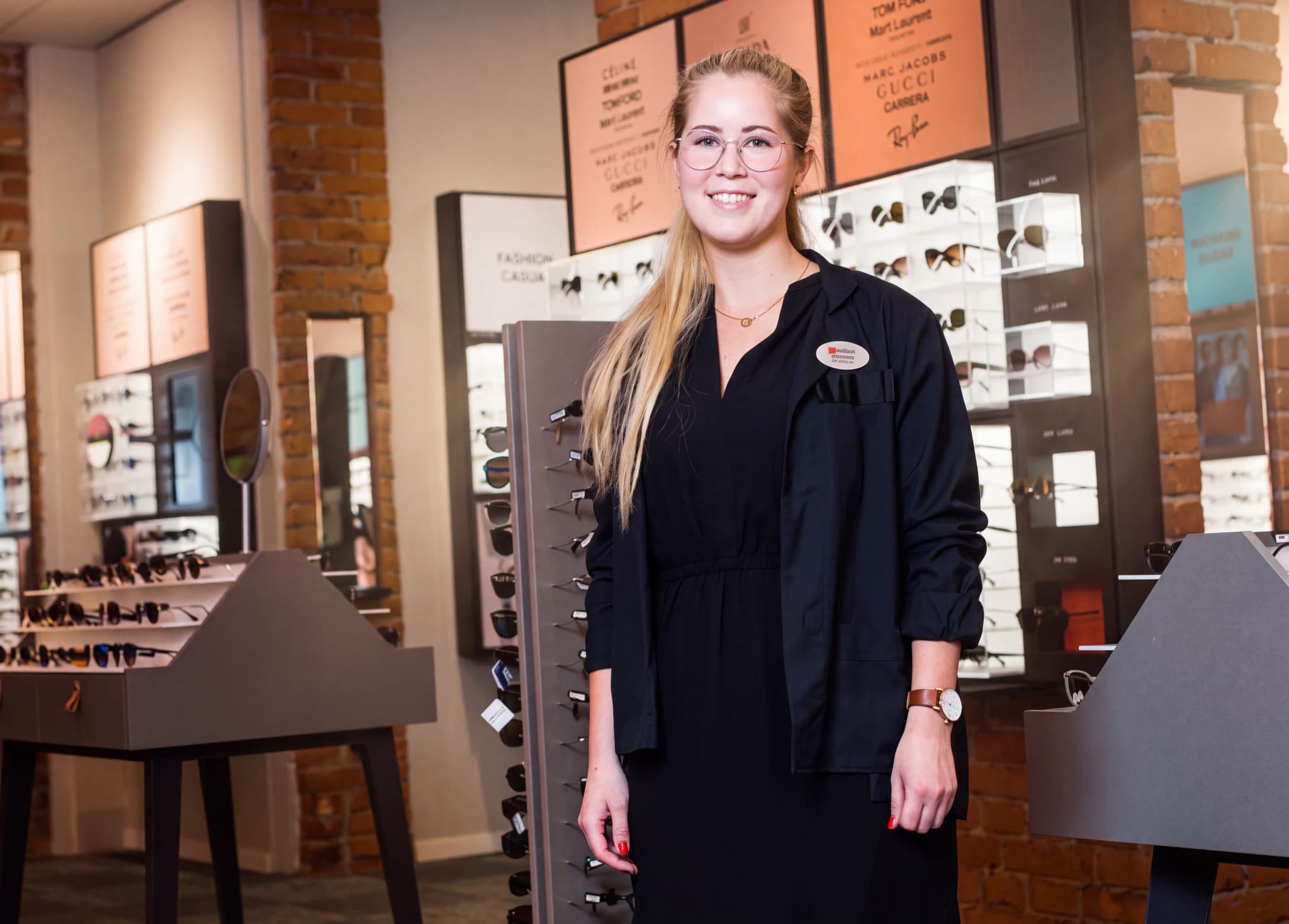 En leende optiker står framför hyllor med glasögon i en optikbutik.