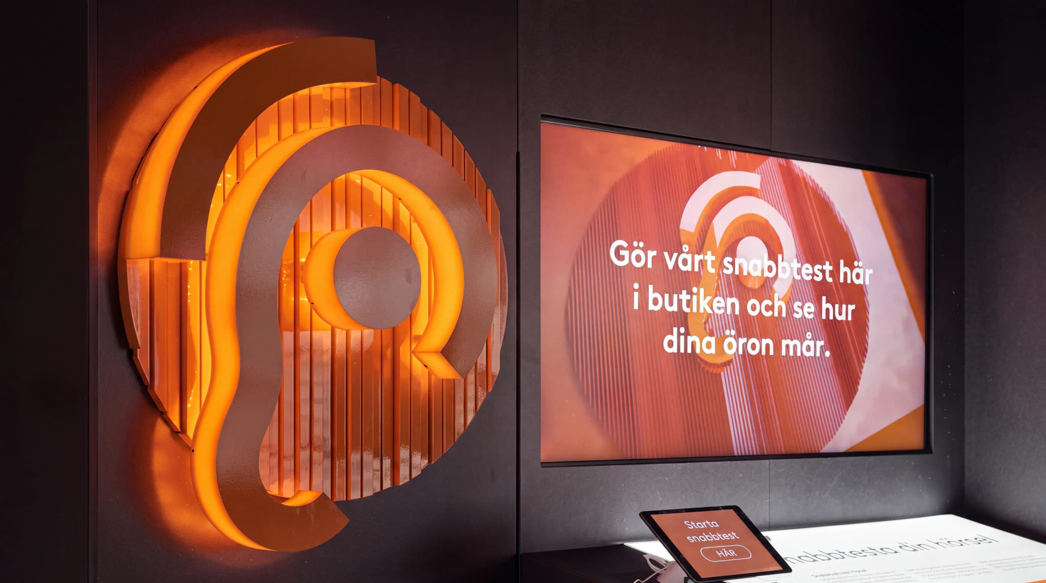 En interaktiv skärm i en butik som visar ett hörseltest med texten "Gör vårt snabbtest här i butiken och se hur dina öron mår.", tillsammans med en stor lysande 3D-logotyp som liknar ett öra.